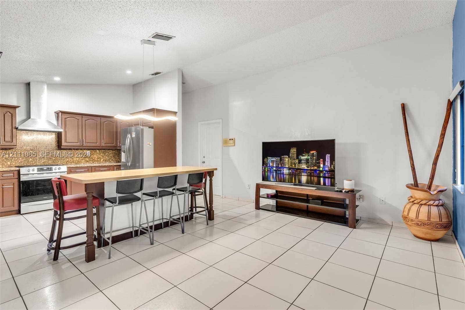 Kitchen featuring wood cabinetry, stainless steel range hood, stone-finish backsplash, and an island with a wood-tone surface and overhead pendant lighting, complemented by tile flooring