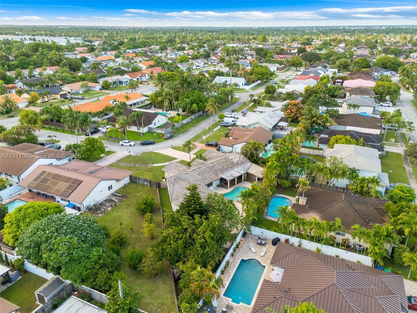 Aerial perspective of a suburban neighborhood featuring residential homes with tile roofs, established tree canopy, and private swimming pools