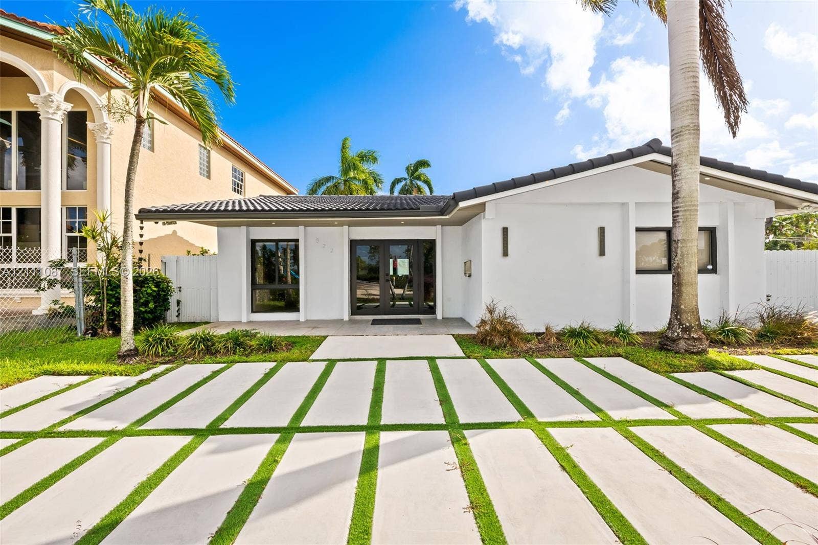822 Polk Street, Hollywood, FL 33019 Single-story exterior featuring white stucco siding, a tiled roof, dual-entry glass doors, and an expansive concrete paver driveway with integrated grass turf