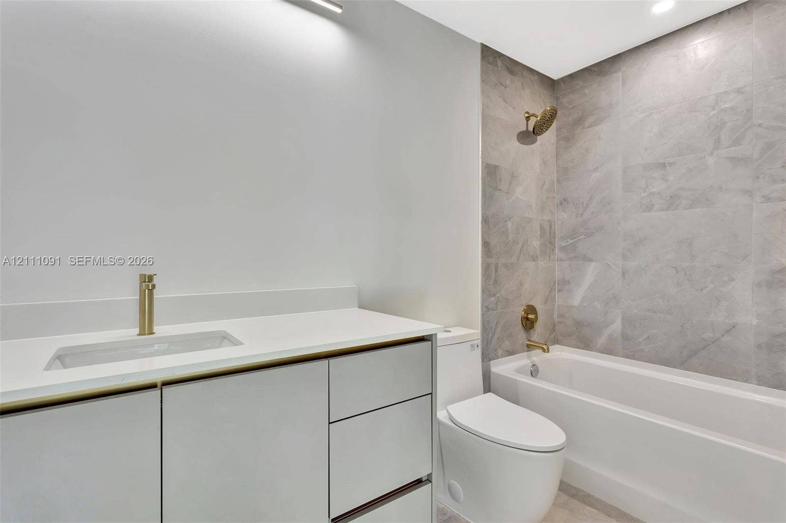 Bathroom featuring a vanity with quartz countertop, light-toned cabinetry, brass-finish fixtures, and a tub enclosure with large-format tile surround