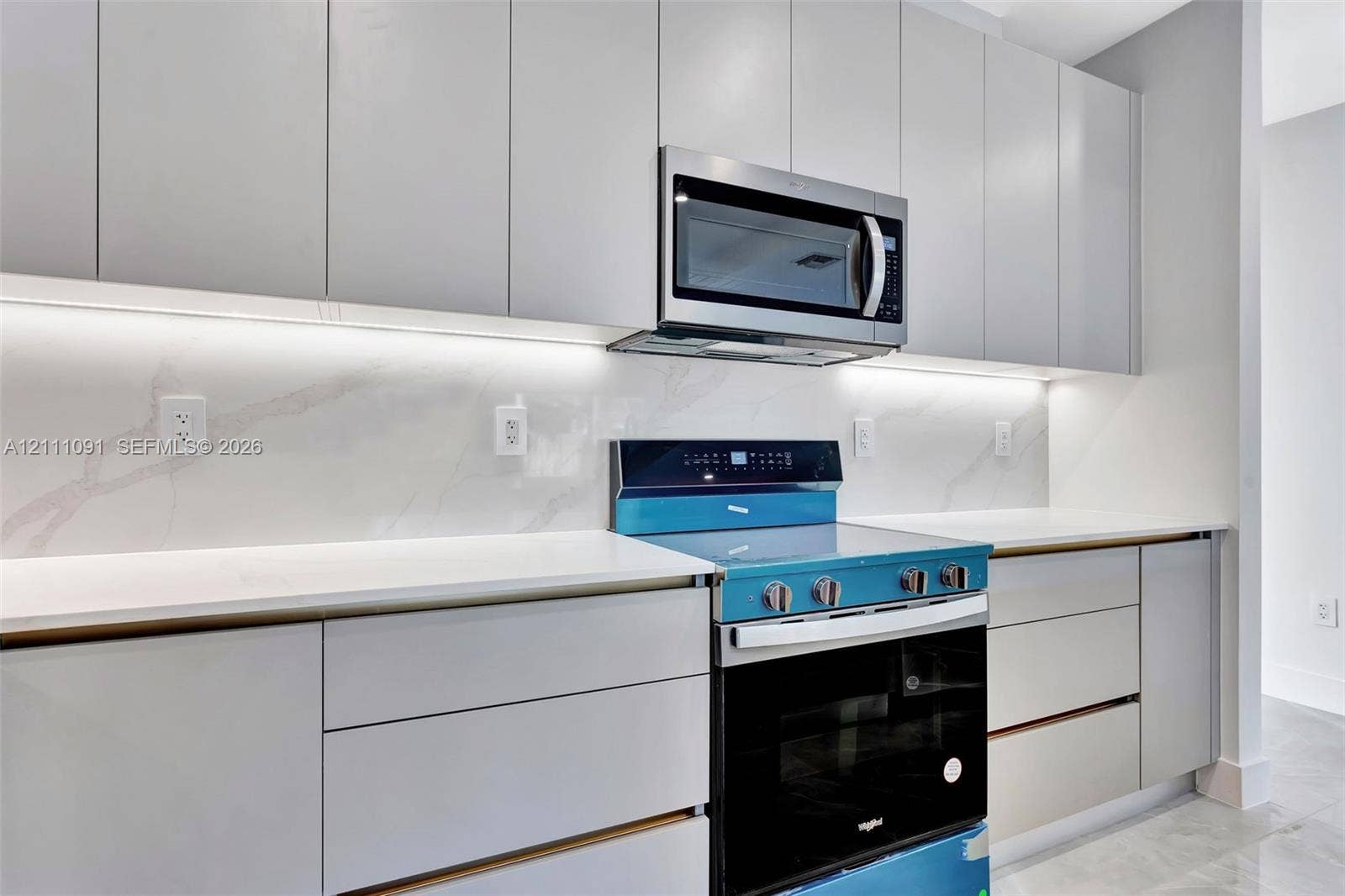 Sleek light-gray flat-panel cabinetry, white stone-finish slab backsplash, integrated under-cabinet lighting, solid-surface countertops, and stainless steel appliances