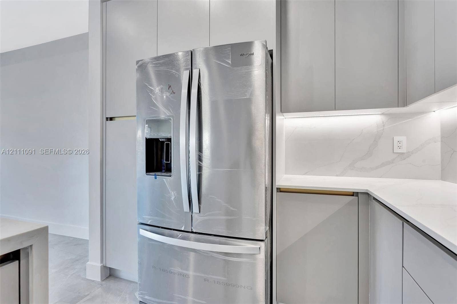 Stainless steel French door refrigerator set within floor-to-ceiling cabinetry, featuring light-toned slab-panel doors, stone-composite countertops, marble-patterned backsplash, and under-cabinet integrated lighting
