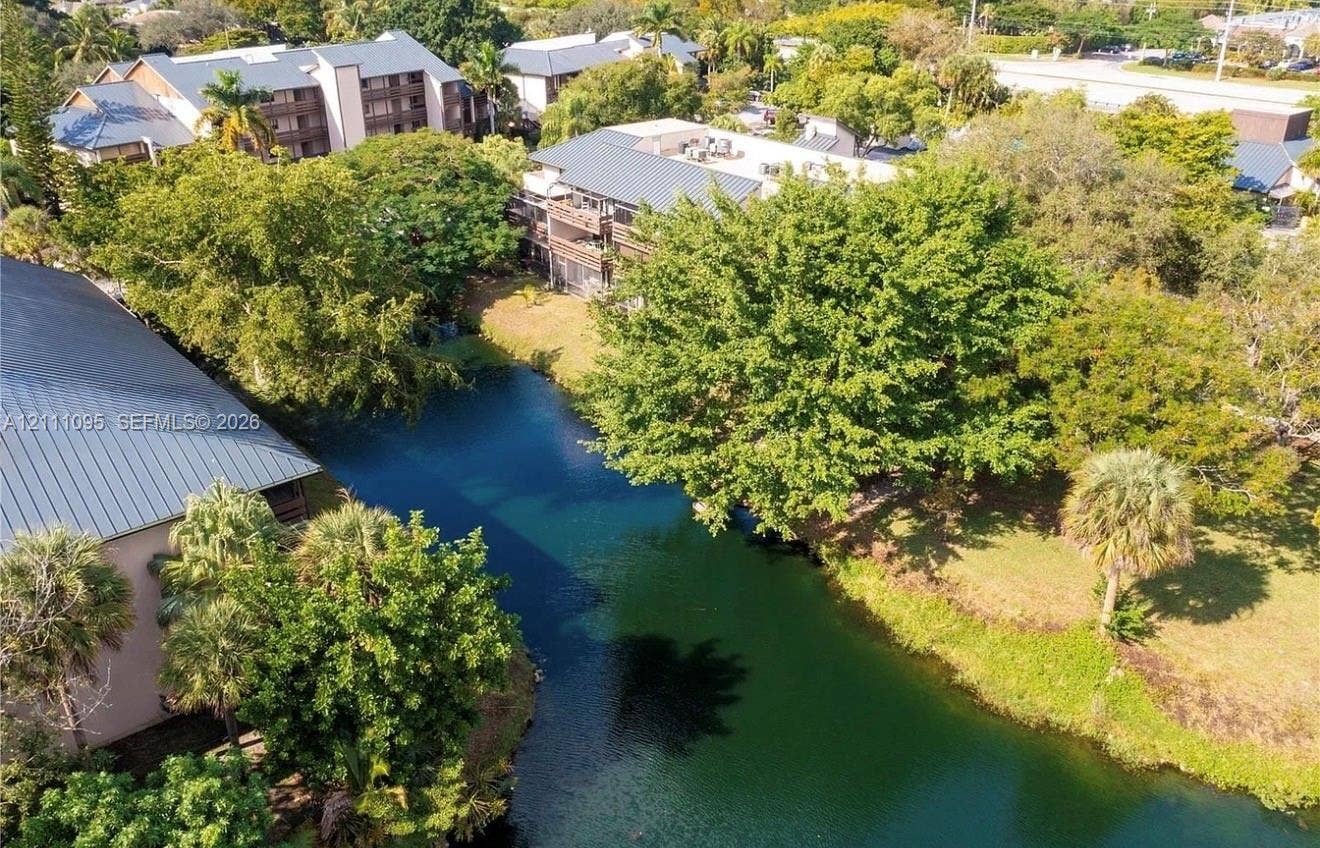 120 SW 96th Terrace, #104, Plantation, FL 33324 Aerial perspective featuring a multi-story building with a metal roof, situated alongside a water feature, accented by mature trees and palm foliage