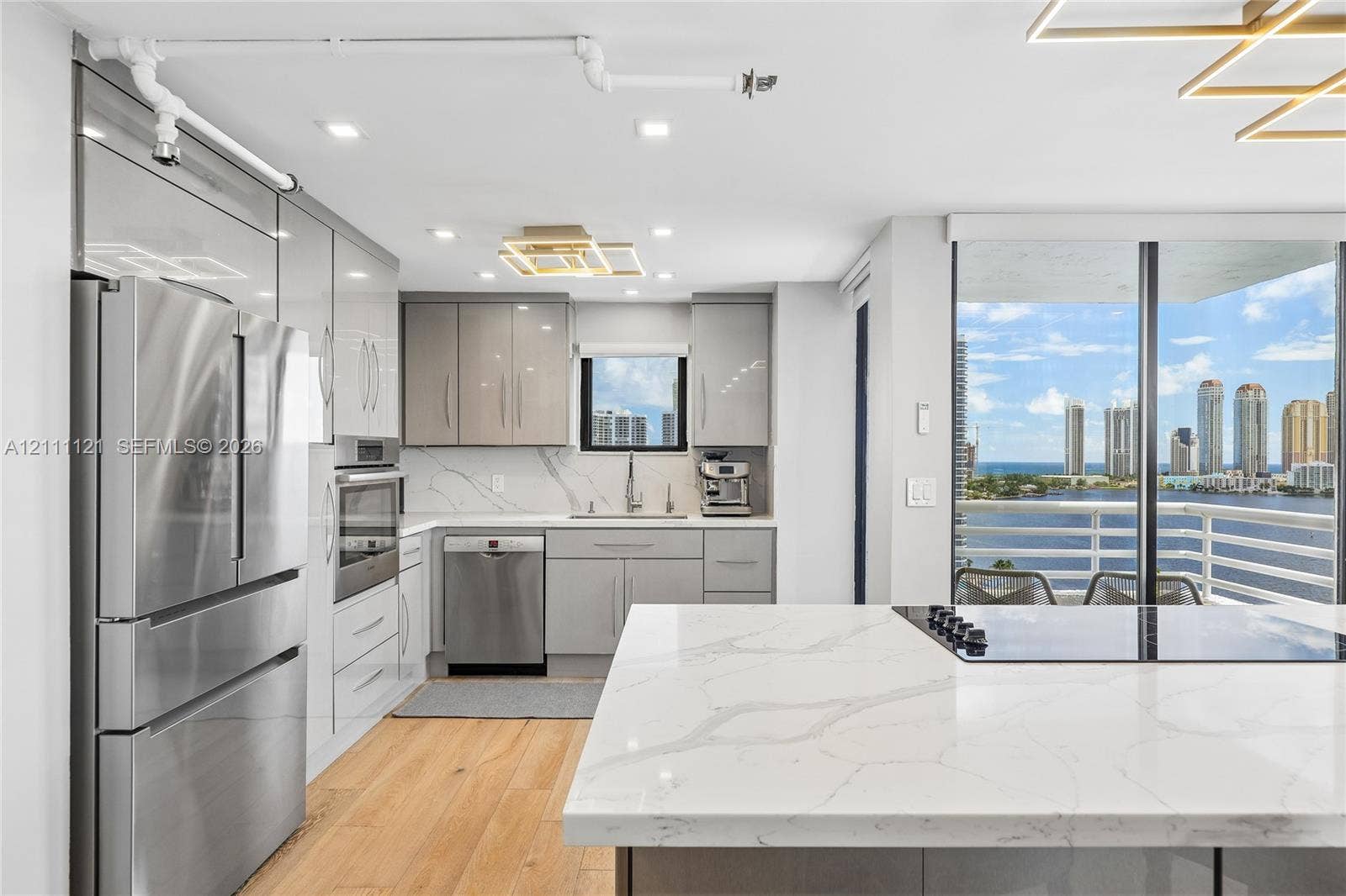 3300 NE 192nd Street, #1813, Aventura, FL 33180 Modern kitchen featuring high-gloss cabinetry, stone-surface countertops with waterfall edge, integrated stainless steel appliances, wood-finish flooring, and floor-to-ceiling glass panel doors