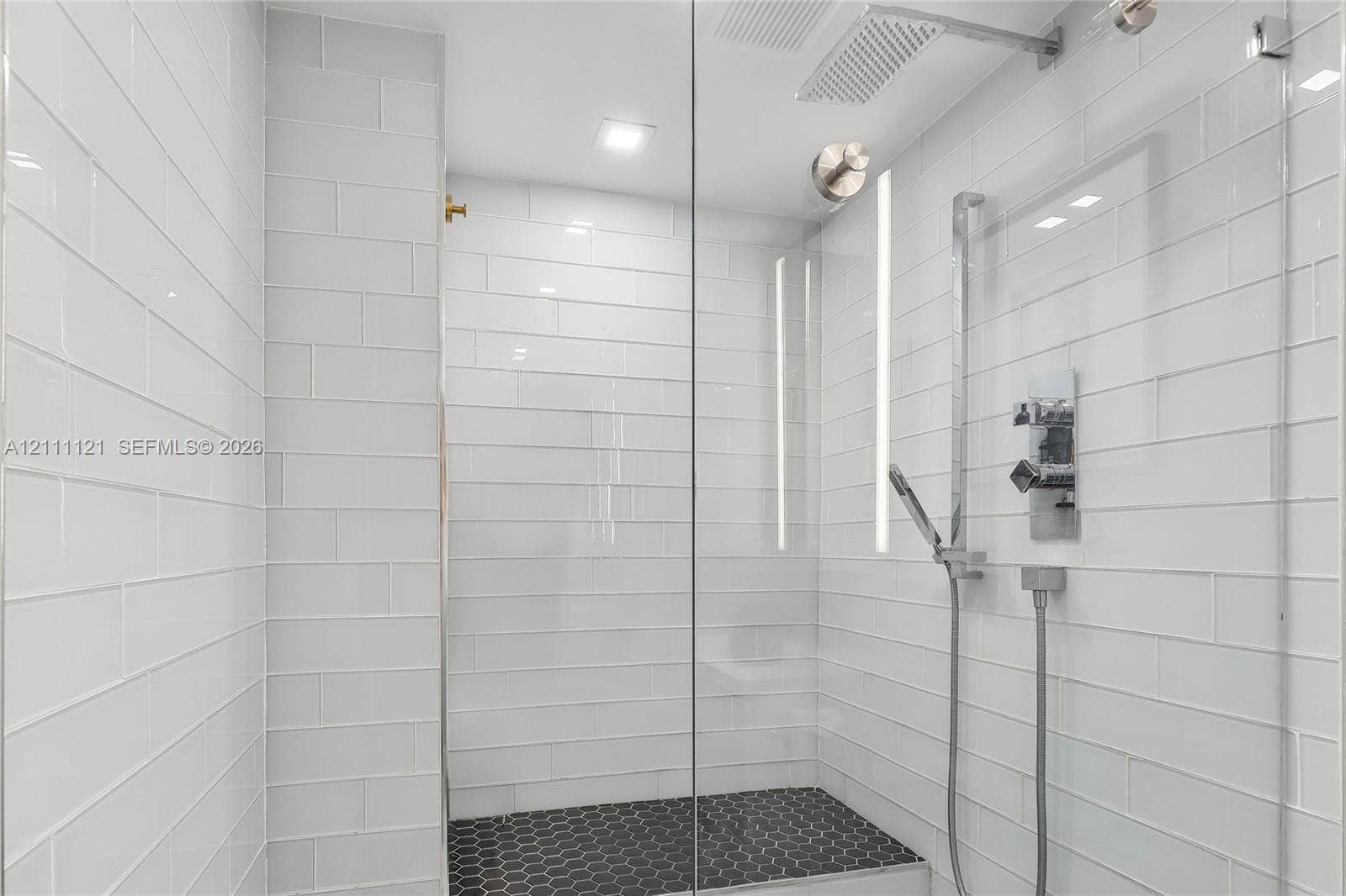 3300 NE 192nd Street, #1813, Aventura, FL 33180 Walk-in shower featuring white subway wall tile, black hexagonal floor tile, a glass enclosure, a rainfall showerhead, and a handheld shower fixture