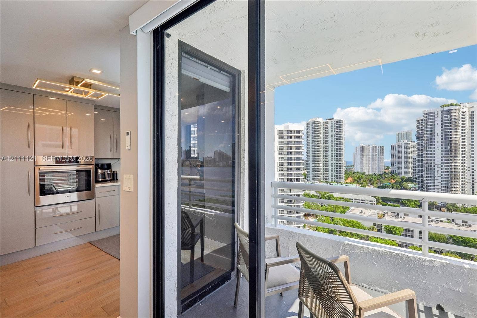 3300 NE 192nd Street, #1813, Aventura, FL 33180 Kitchen featuring high-gloss cabinetry, stainless steel built-in wall oven, wood-finish flooring, and a balcony with a white metal railing and a cityscape backdrop