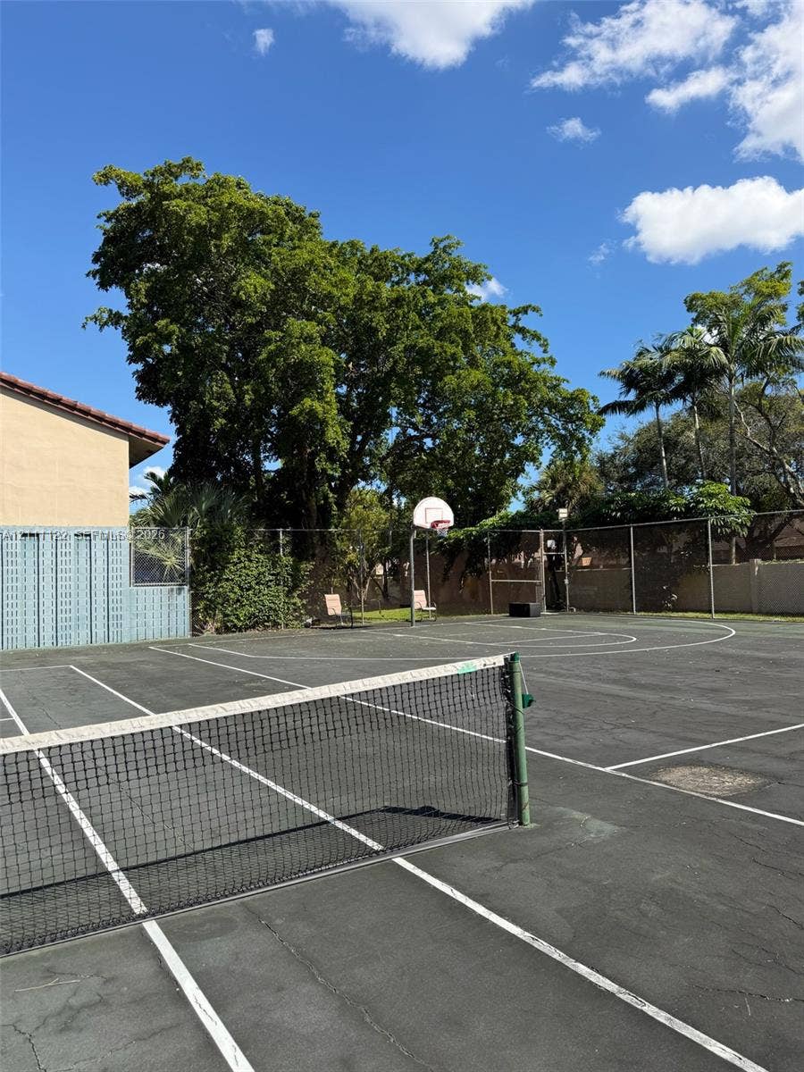Multi-purpose sport court featuring a tennis net, basketball hoop, perimeter fencing, and mature trees