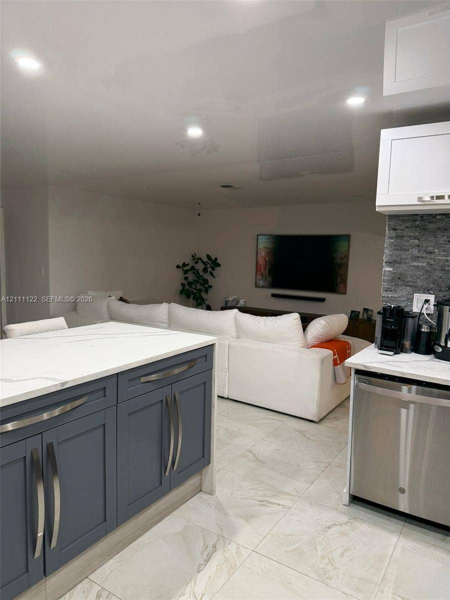 Kitchen featuring a center island with a white-veined countertop, slate-blue cabinetry, stainless steel dishwasher, stone-tiled backsplash, and large-format porcelain tile flooring
