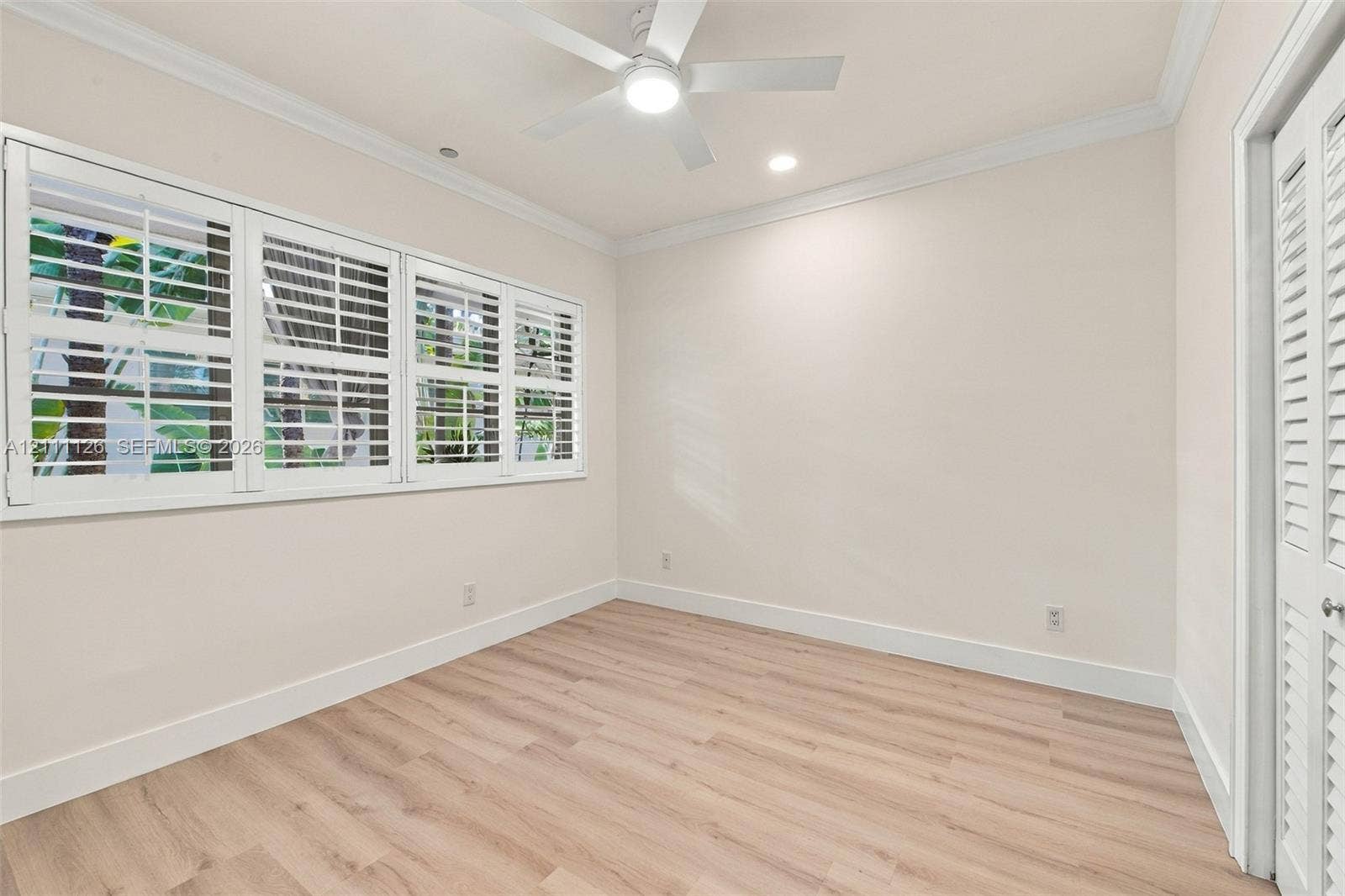 Light-filled room featuring wood-finish flooring, plantation shutters, crown molding, a central ceiling fan with integrated lighting, and louvered closet doors