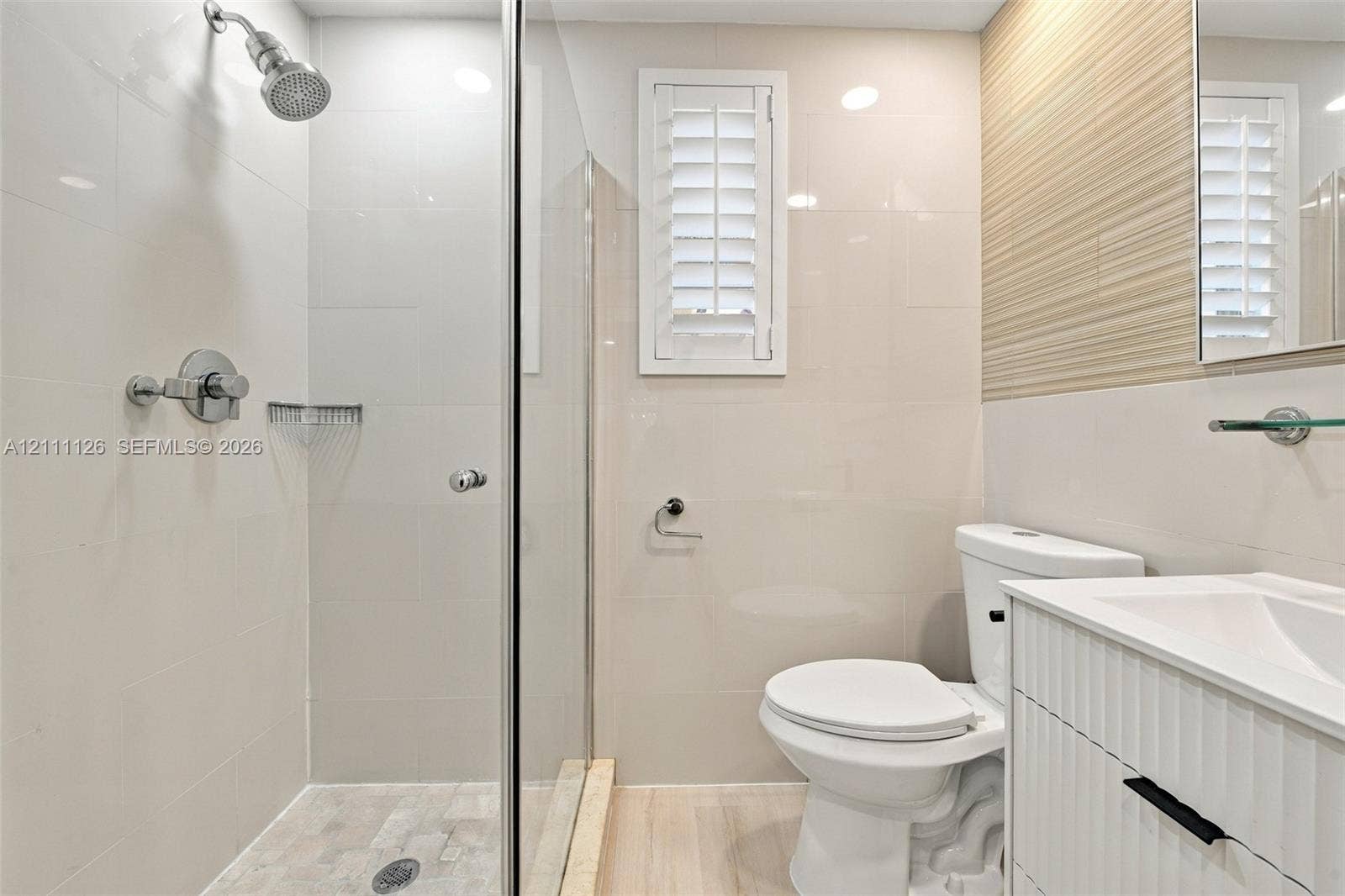 Walk-in shower with glass enclosure, wall-mounted shower head, textured accent wall, integrated vanity with white cabinetry, and shutter-style window treatment