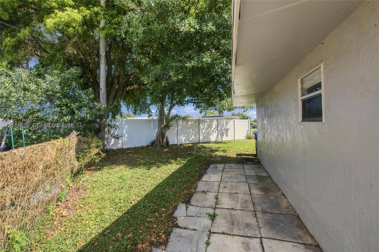 Side yard featuring a concrete paver walkway, white vinyl privacy fencing, chain-link perimeter fencing, and mature trees