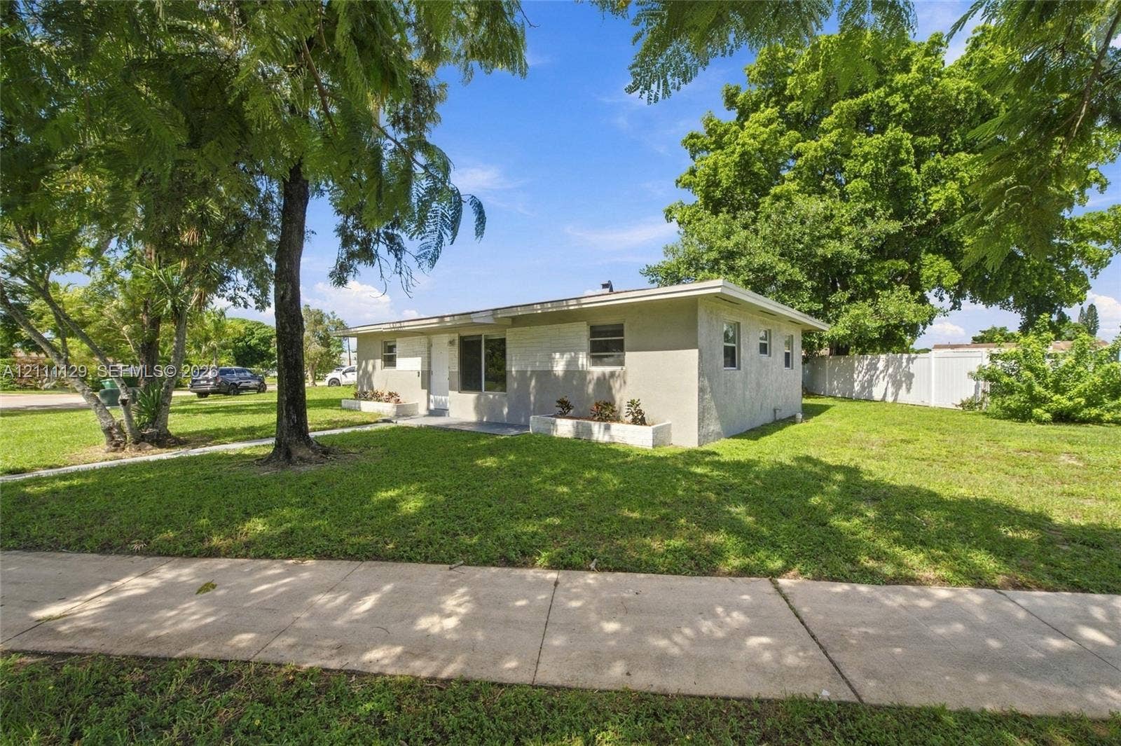 Single-story residence featuring a light-toned stucco exterior, integrated concrete planters, and mature shade trees on a corner lot with white vinyl fencing