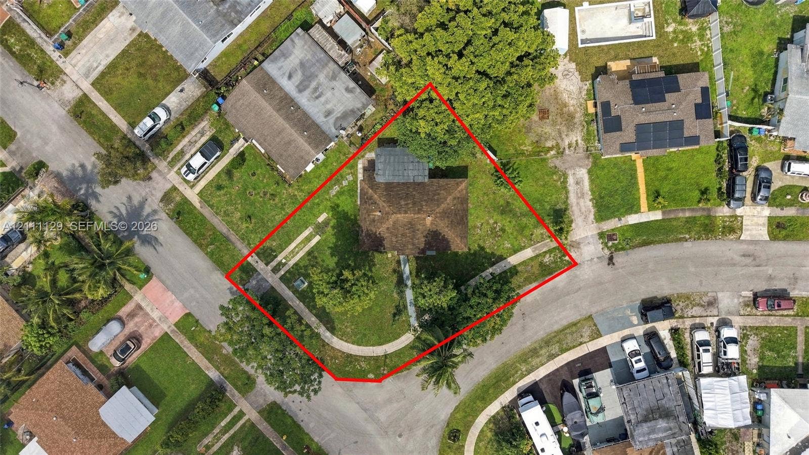 Aerial perspective of a corner lot featuring a hipped roof structure, curved concrete walkway, mature trees, and ample green space