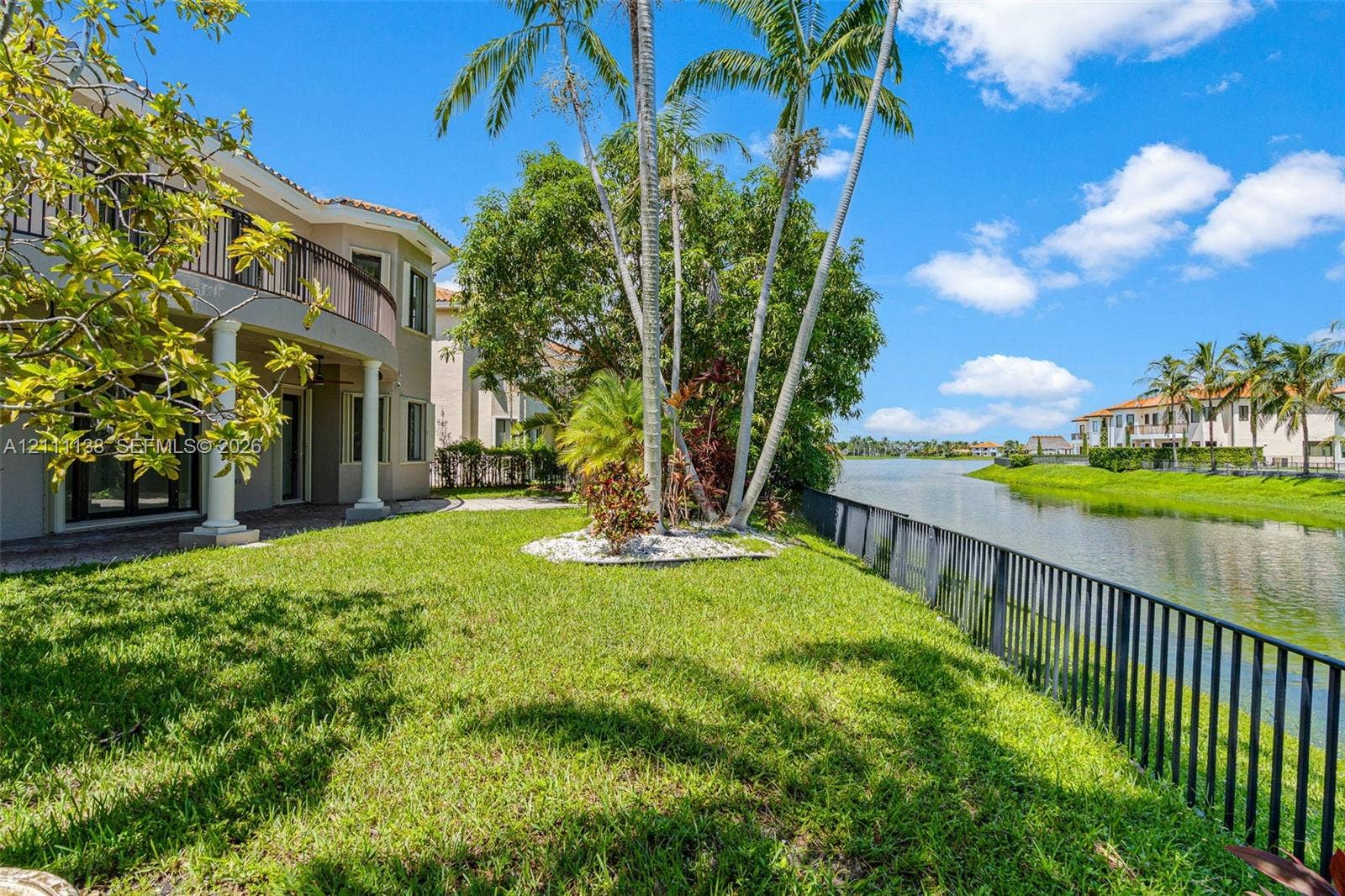 19363 SW 79th Avenue, Cutler Bay, FL 33157 Waterfront backyard featuring a manicured lawn, metal perimeter fencing, mature palm trees, and a two-story exterior with a curved balcony
