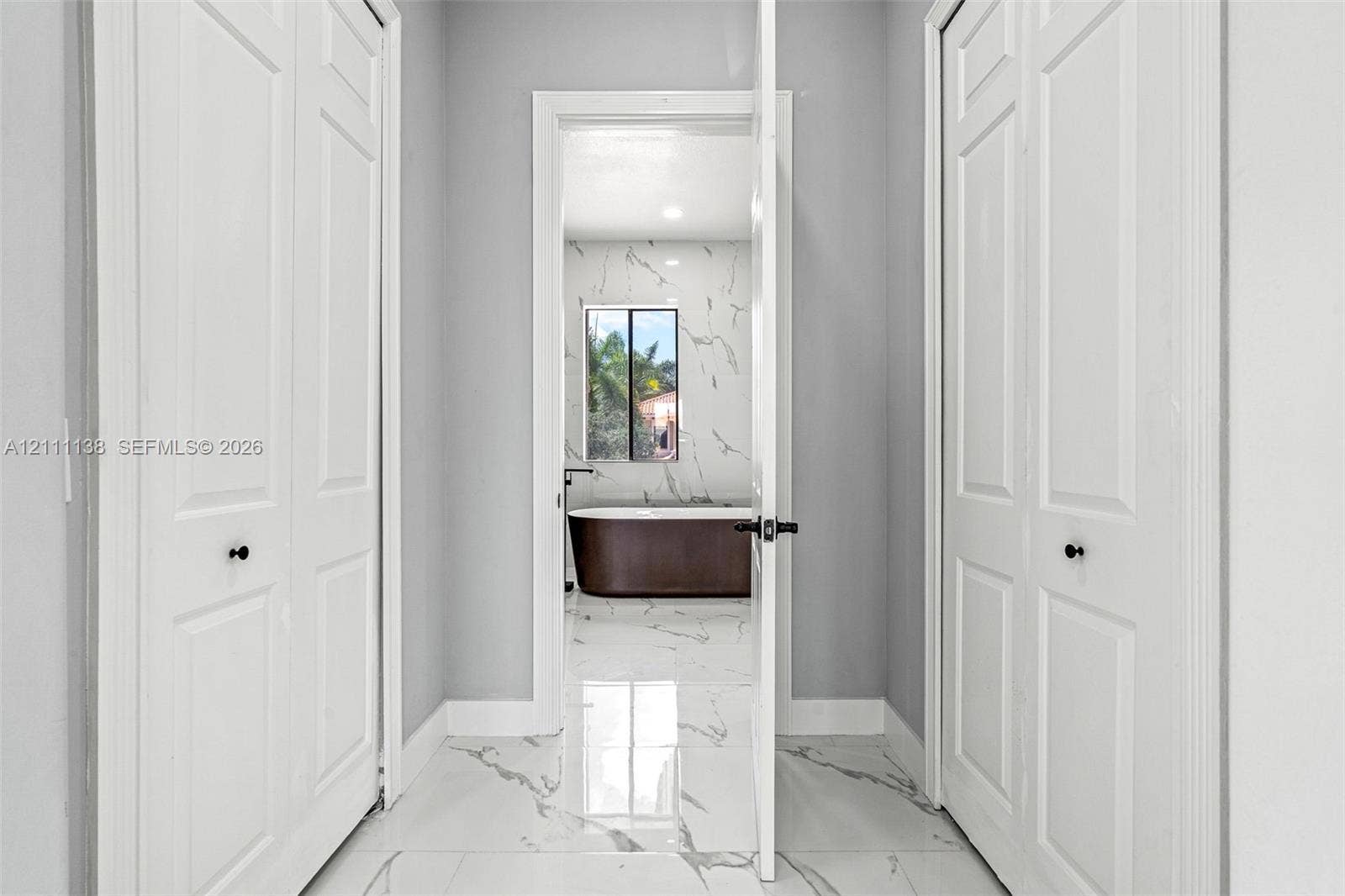 19363 SW 79th Avenue, Cutler Bay, FL 33157 Hallway featuring white panel doors with matte black hardware and polished stone-finish floor tile with veining, leading into a space with matching stone-finish wall tile and a freestanding soaking tub