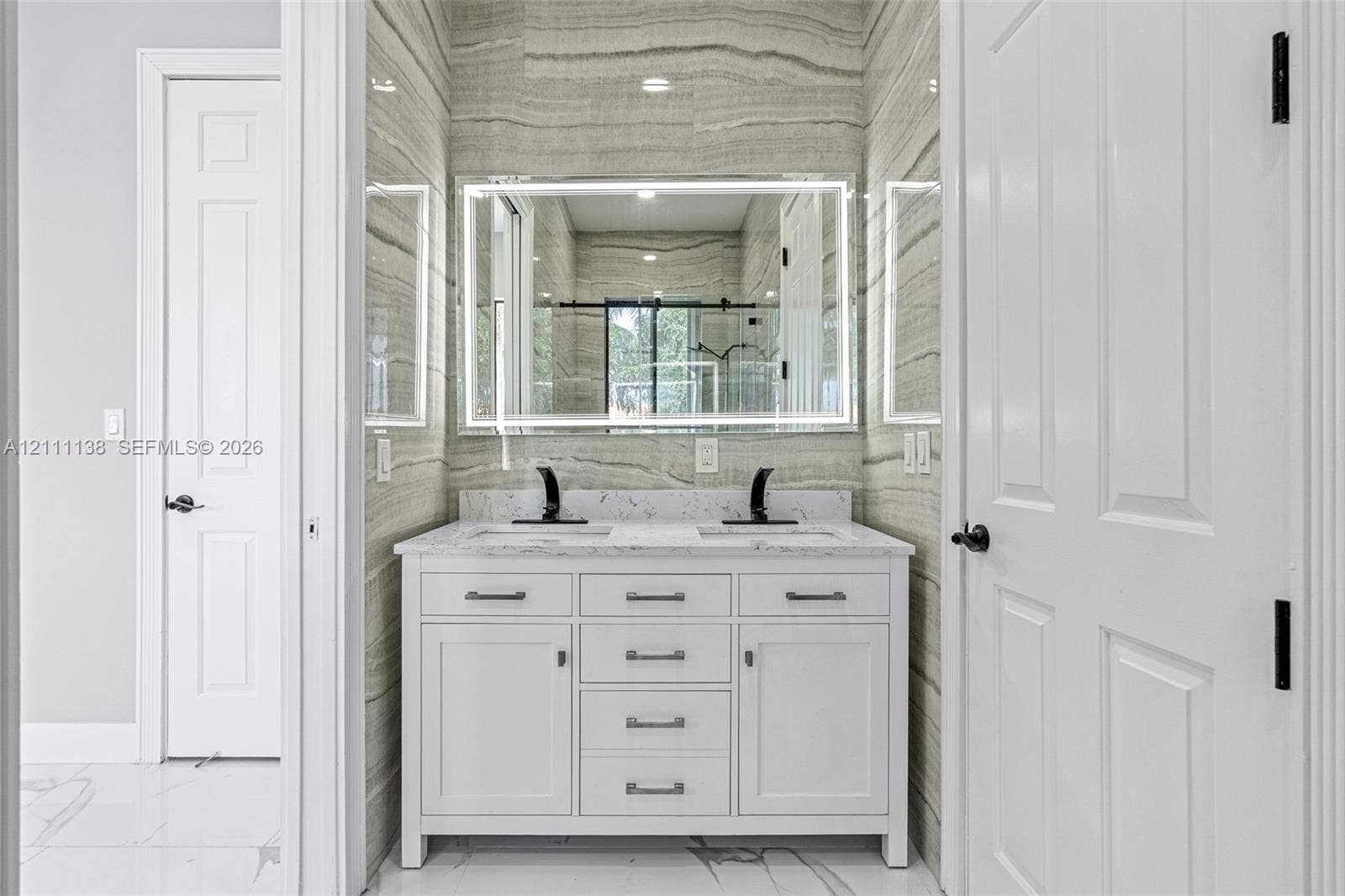 19363 SW 79th Avenue, Cutler Bay, FL 33157 Double vanity featuring a white cabinetry base, stone-look countertop, integrated dual sinks, and matte black widespread faucets, accented by full-height linear pattern tile walls and a modern LED-backlit mirror