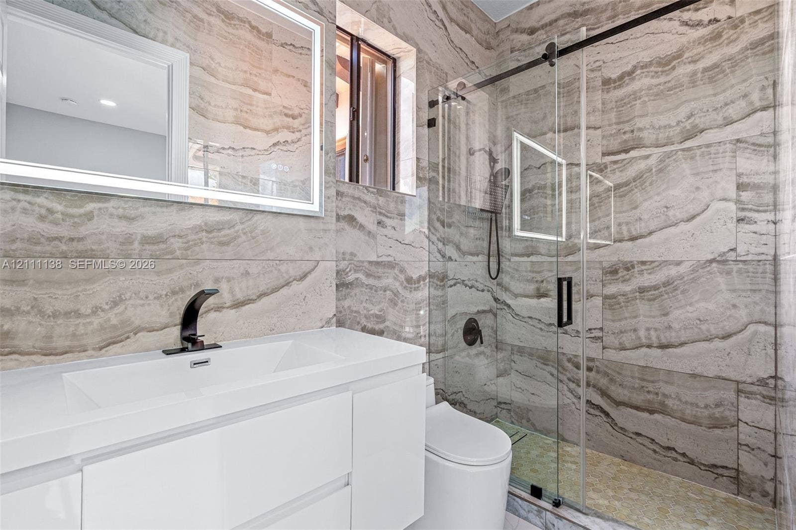 19363 SW 79th Avenue, Cutler Bay, FL 33157 Modern vanity with a rectangular integrated sink, wall-mounted backlit mirror, walk-in shower with sliding glass door, and large-format natural stone wall tiling