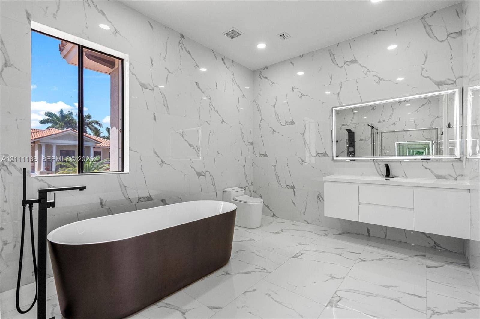 19363 SW 79th Avenue, Cutler Bay, FL 33157 Bathroom featuring floor-to-ceiling large-format tile with stone-pattern finish, wall-mounted floating vanity, freestanding soaking tub with matte black floor-mounted faucet, and an oversized LED-backlit mirror
