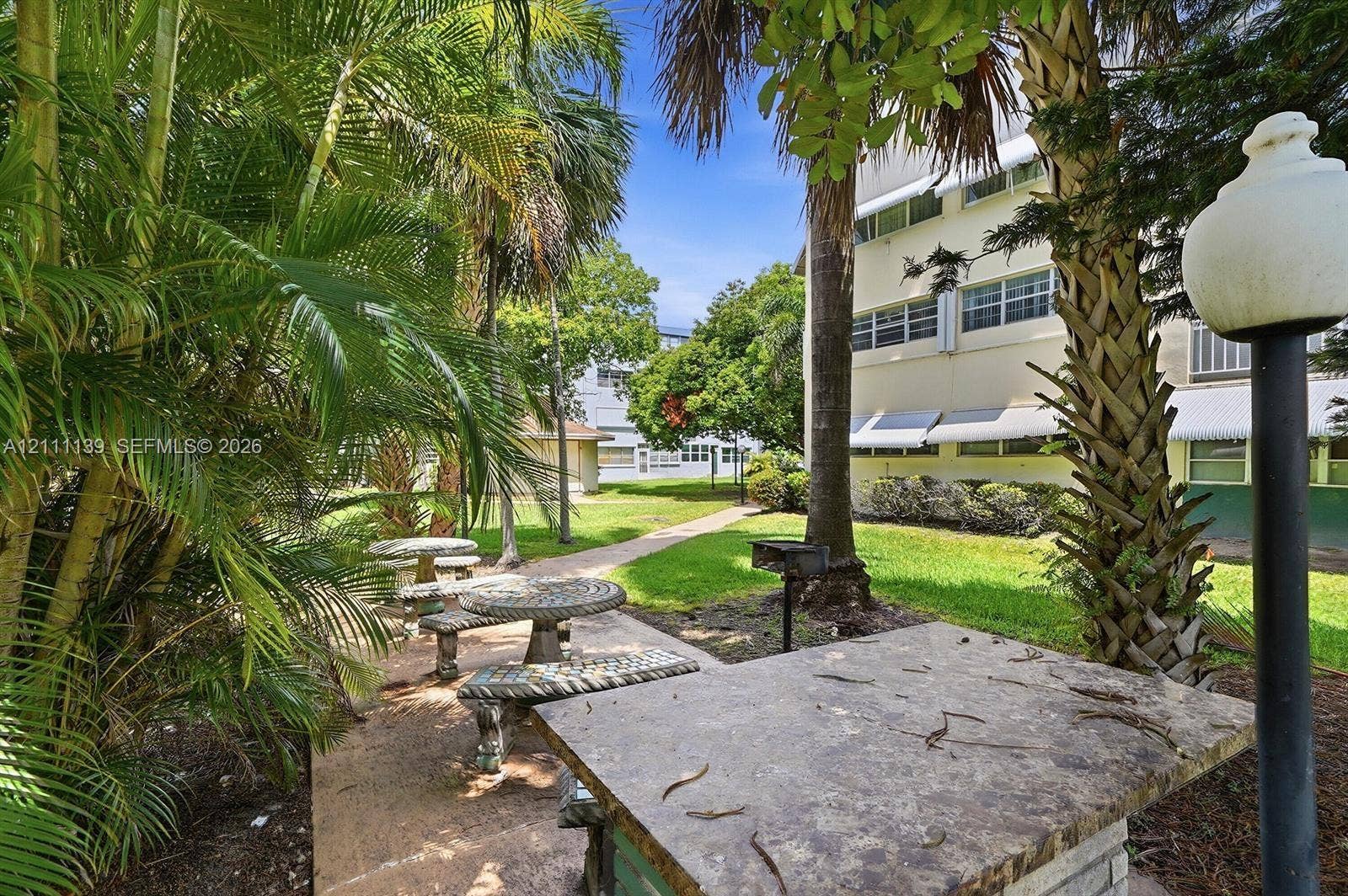 1333 E Hallandale Beach Boulevard, #236, Hallandale Beach, Hallandale Beach, FL 33009 Common area featuring stone-finish picnic tables, a built-in outdoor grill, manicured lawn, and mature palm trees