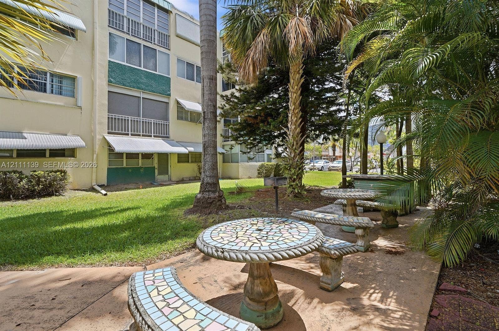 1333 E Hallandale Beach Boulevard, #236, Hallandale Beach, Hallandale Beach, FL 33009 Common courtyard featuring a manicured lawn, mature palm trees, and decorative stone-tiled picnic tables with matching benches