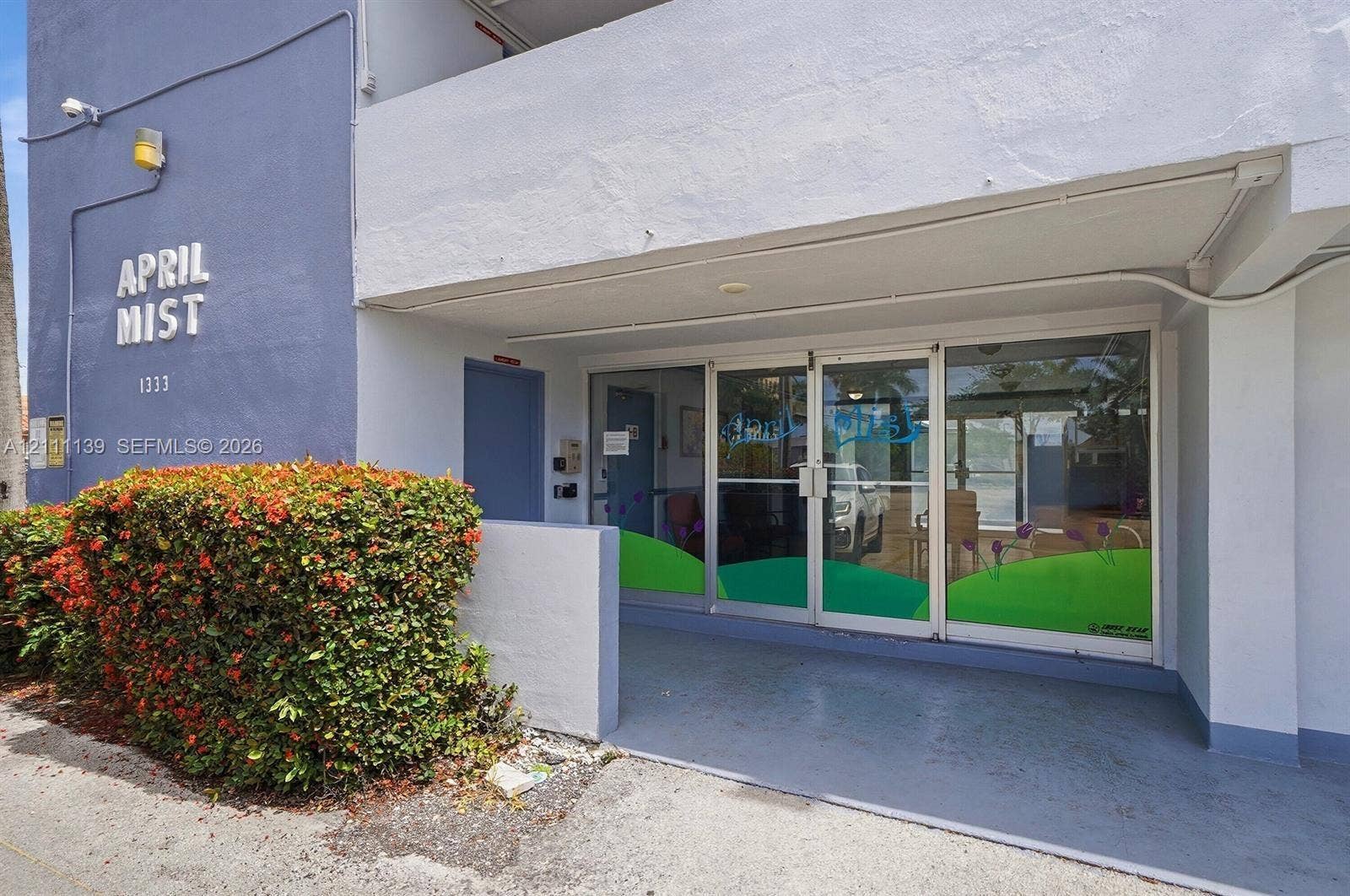 1333 E Hallandale Beach Boulevard, #236, Hallandale Beach, Hallandale Beach, FL 33009 Exterior building entrance featuring a concrete facade, aluminum-framed glass storefront doors, a covered porch, and mature landscaping