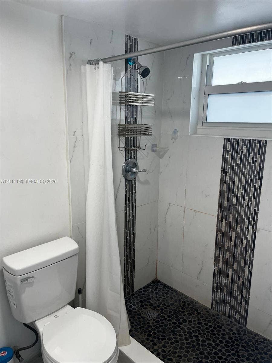 1333 E Hallandale Beach Boulevard, #236, Hallandale Beach, Hallandale Beach, FL 33009 Shower featuring white wall tile with vertical mosaic accent inlay, pebble-style flooring, and a single-handle chrome fixture, paired with a standard-height porcelain toilet