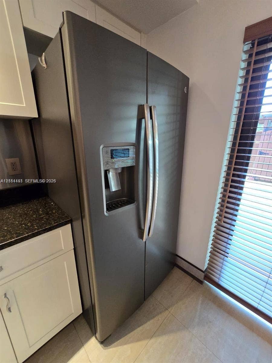 Stainless steel side-by-side refrigerator with exterior water and ice dispenser, dark stone countertops, white shaker-style cabinetry, and horizontal window blinds