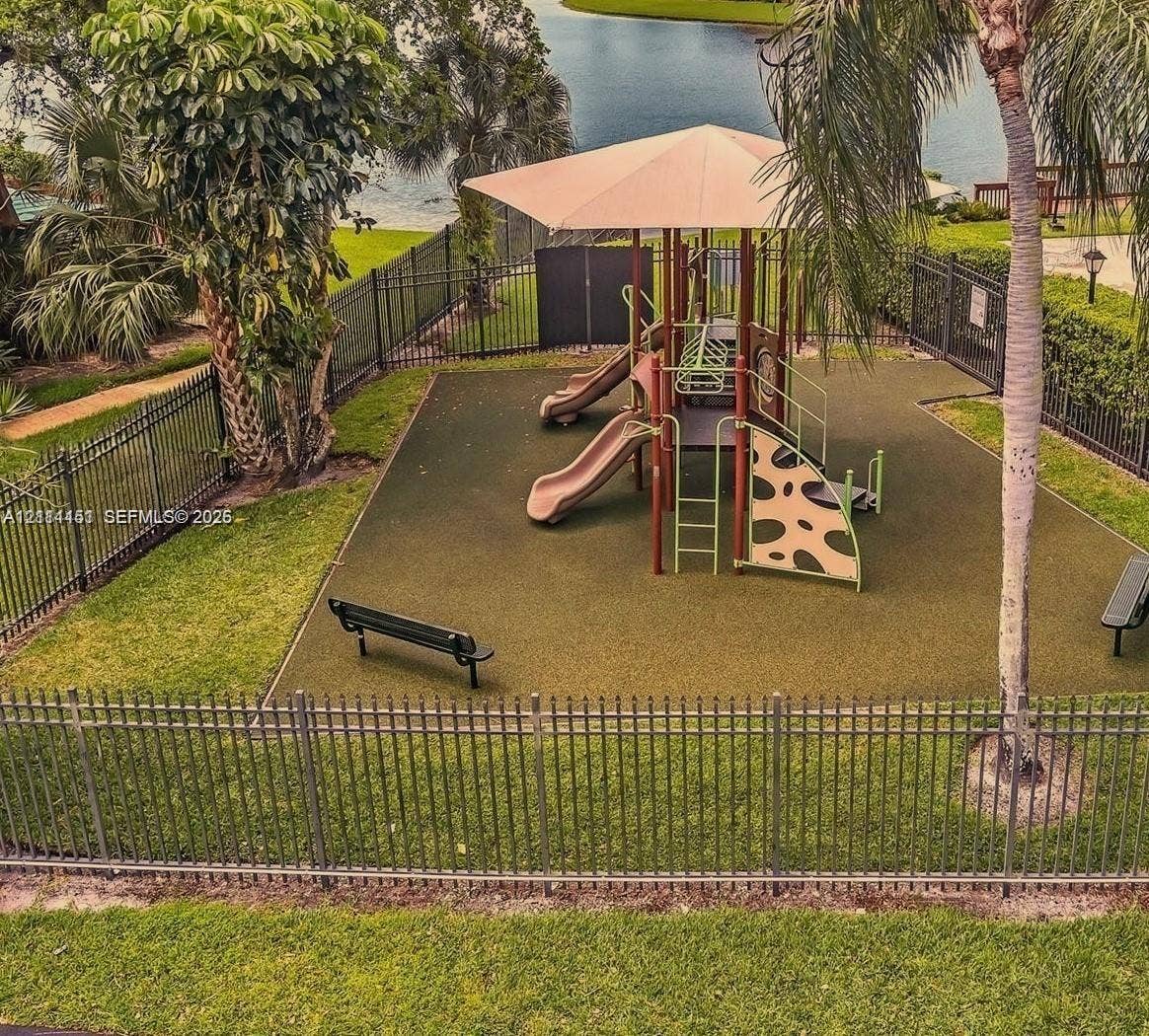 Playground area featuring a multi-level structure with slides, a tensioned shade canopy, rubberized surfacing, and black metal perimeter fencing situated on the waterfront