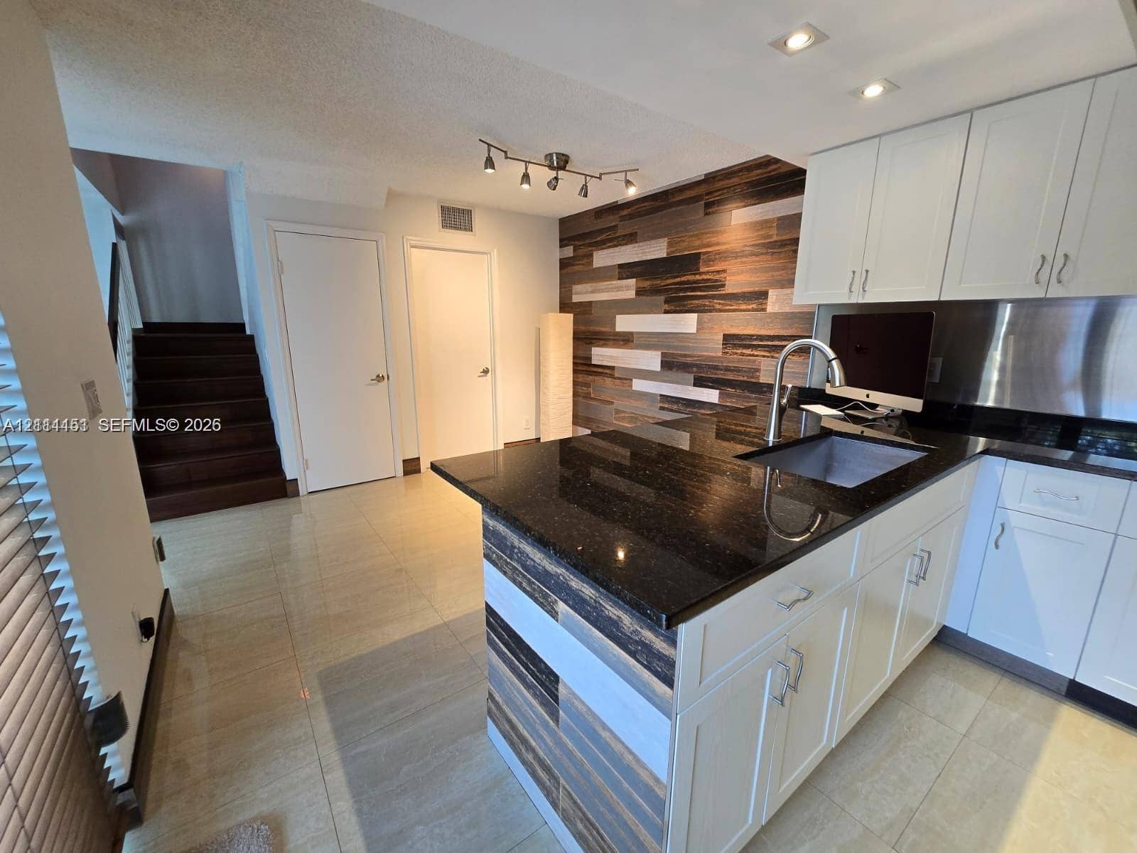 Kitchen island featuring a dark stone countertop, undermount sink, and multi-toned wood-finish paneling, complemented by white cabinetry, stainless steel backsplash, and a decorative wood-finish wall accent