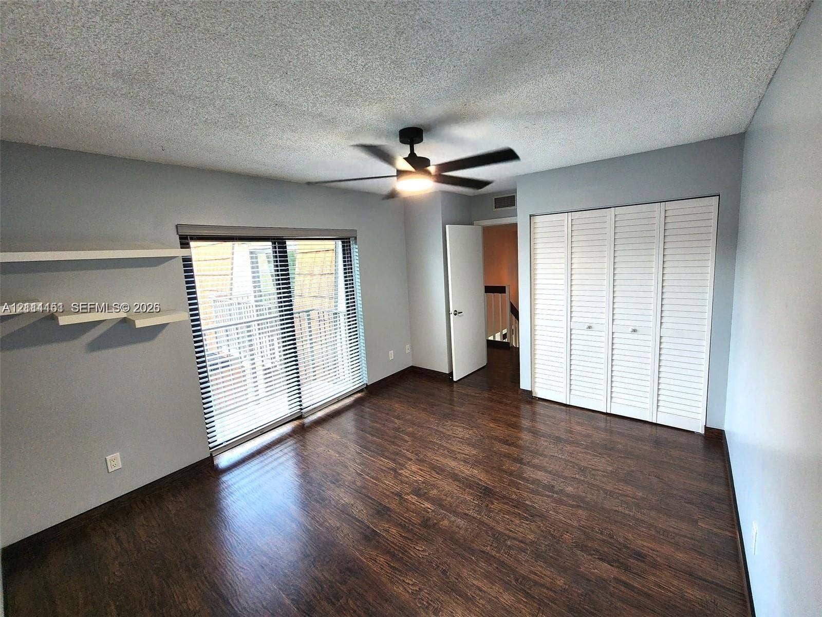 Room featuring wood-finish flooring, sliding glass door with horizontal blinds, built-in wall shelving, multi-panel louvered closet doors, and a ceiling fan with light fixture