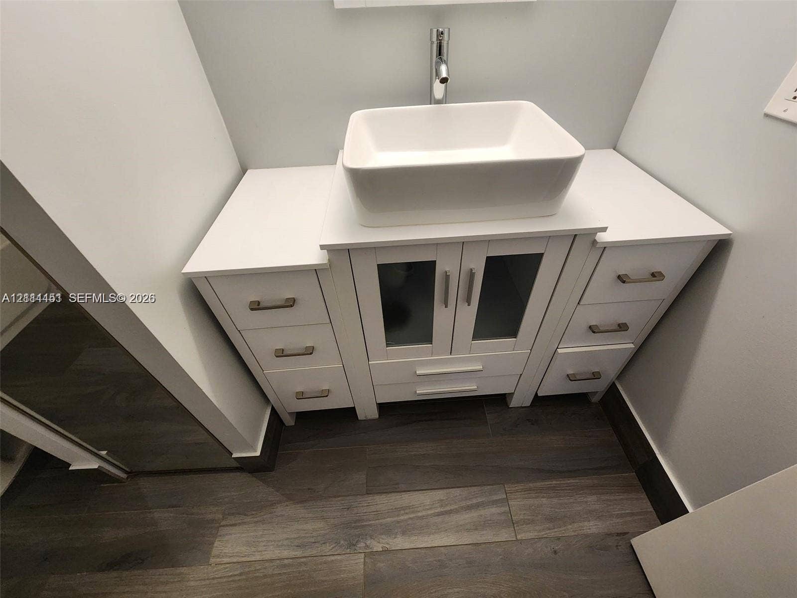 White vanity featuring a rectangular vessel sink, solid surface countertop, glass-panel cabinet doors, brushed metal hardware, and wood-finish flooring