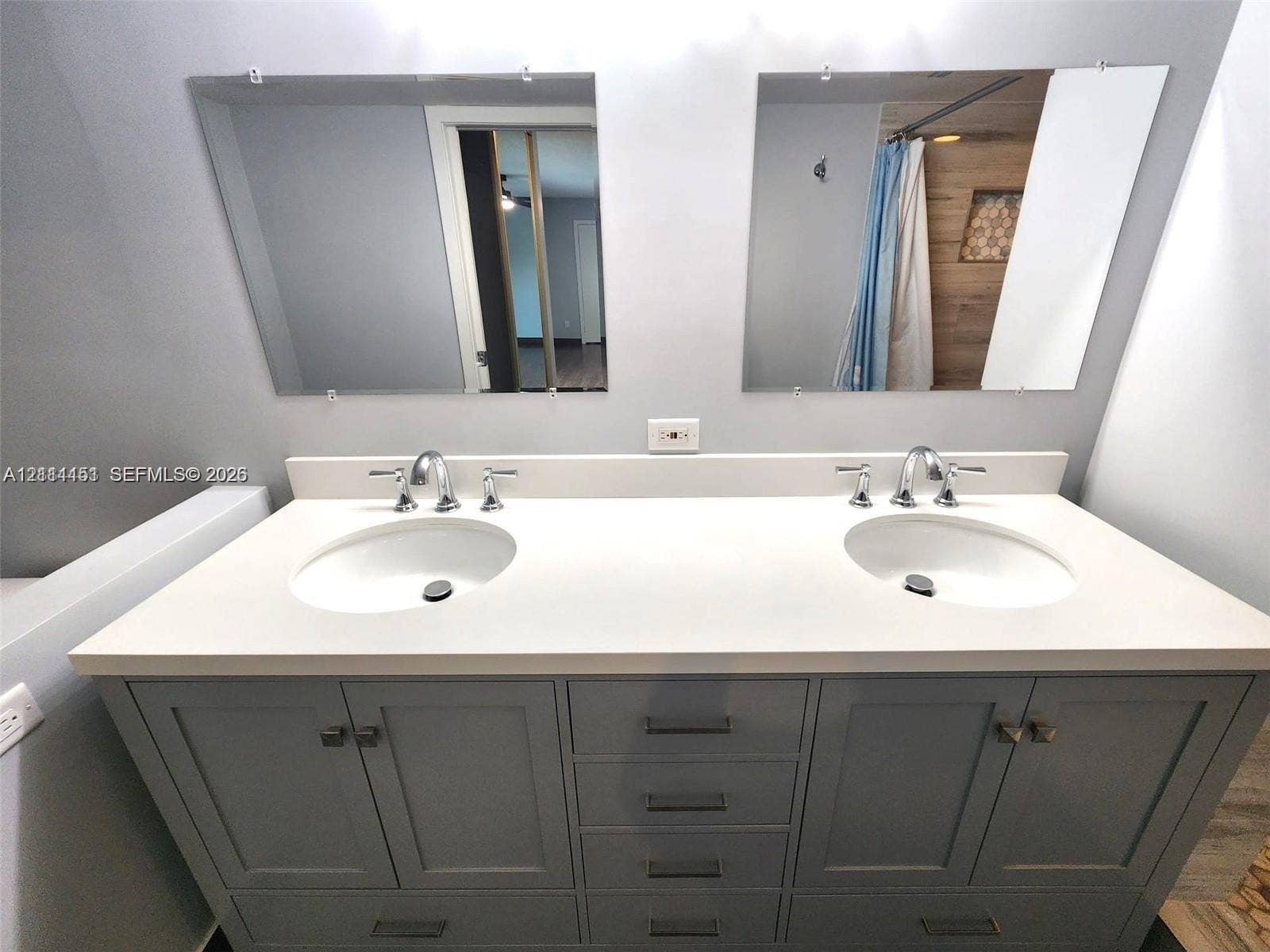 Double vanity featuring a light-toned solid surface countertop, dual undermount sinks, chrome widespread faucets, and gray Shaker-style cabinetry with matte finish hardware, topped by two frameless wall-mounted mirrors