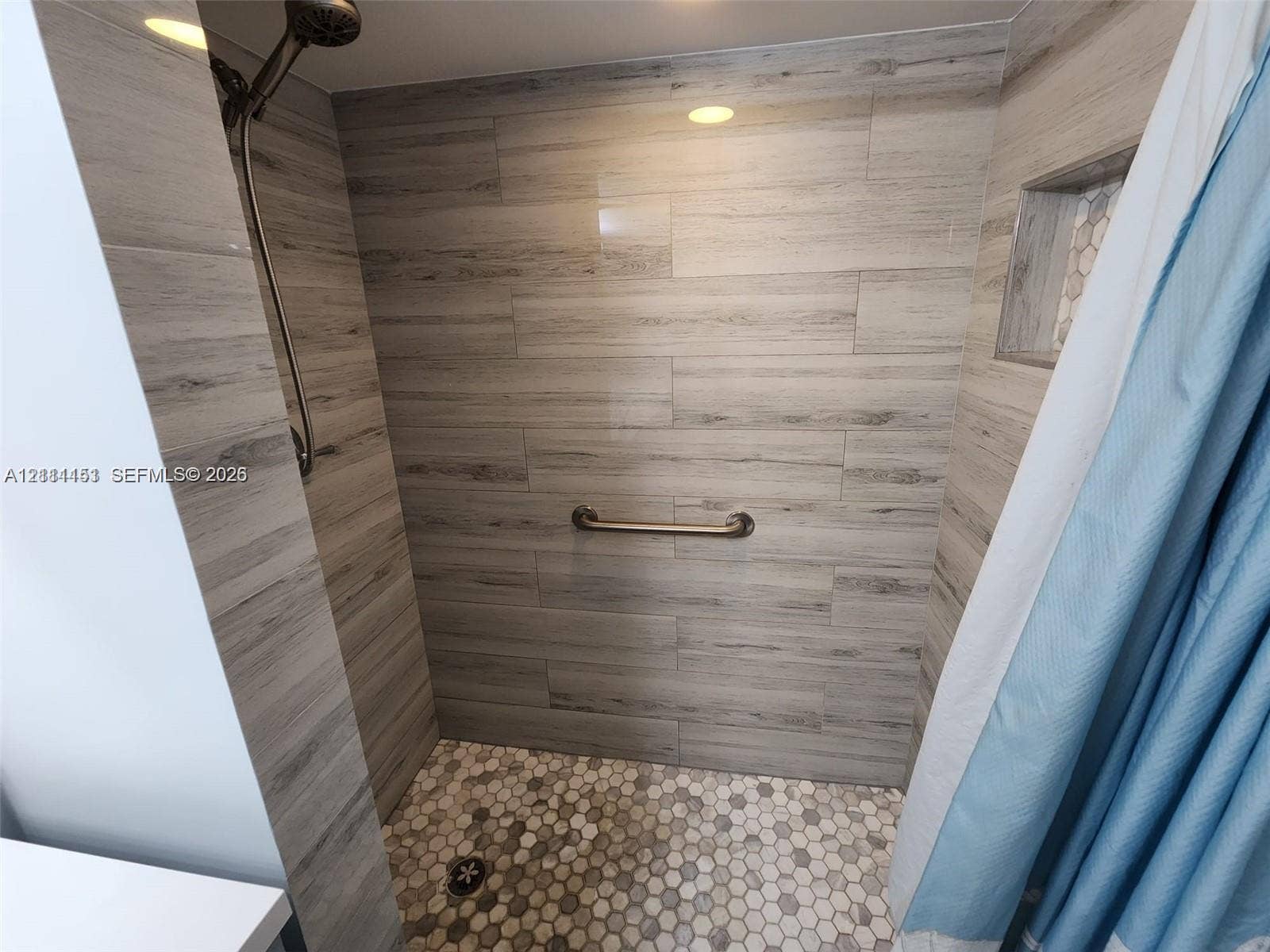 Walk-in shower with wood-finish tile wall surround, hexagonal floor tiling, recessed wall niche, stainless steel shower head, and wall-mounted grab bar