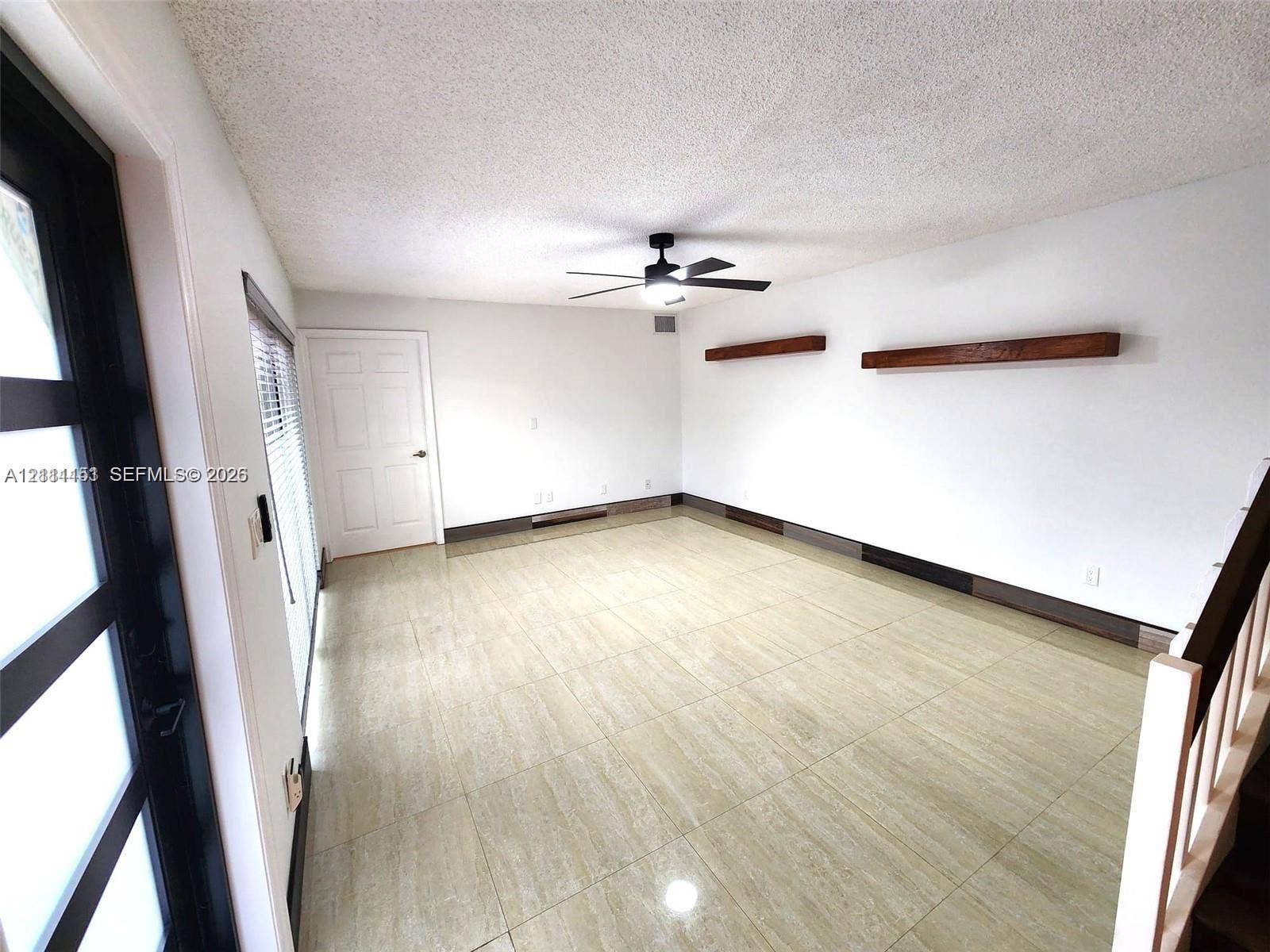 Neutral-toned tile flooring, dual wooden floating shelves, black-framed glass entry door, and a central ceiling fan with integrated light fixture