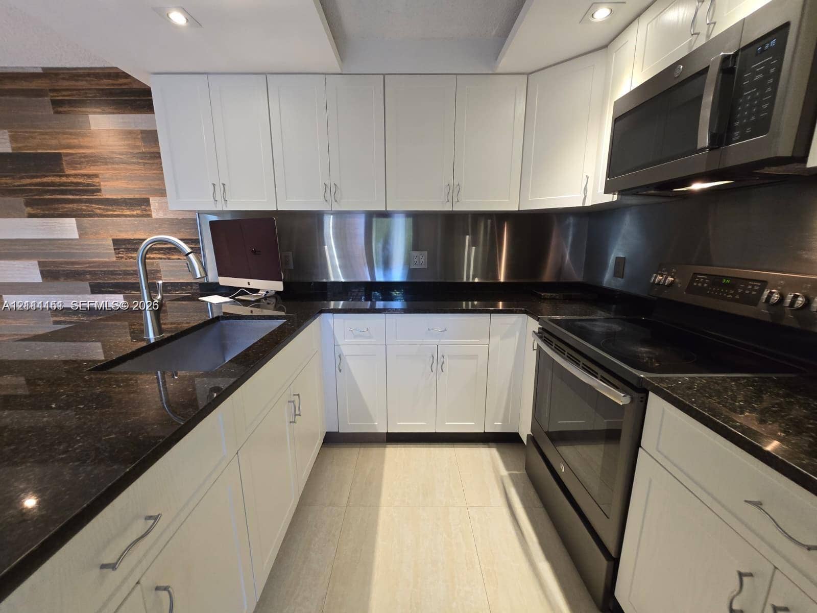 Kitchen featuring white shaker-style cabinetry, dark stone countertops, stainless steel backsplash, integrated appliances, and wood-finish accent wall paneling