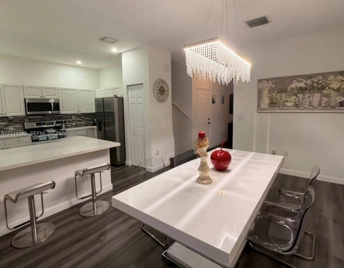 Kitchen featuring white cabinetry, a breakfast bar with stone-finish countertop, stainless steel appliances, a multi-tonal linear tile backsplash, and a rectangular crystal-accented chandelier