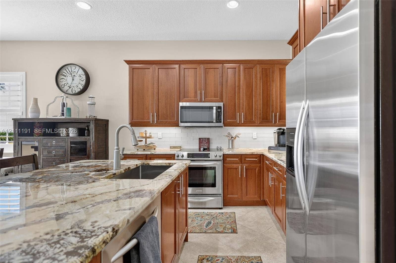 Kitchen featuring wood cabinetry, stone countertops, stainless steel appliances, a center island with an undermount sink, and tile backsplash