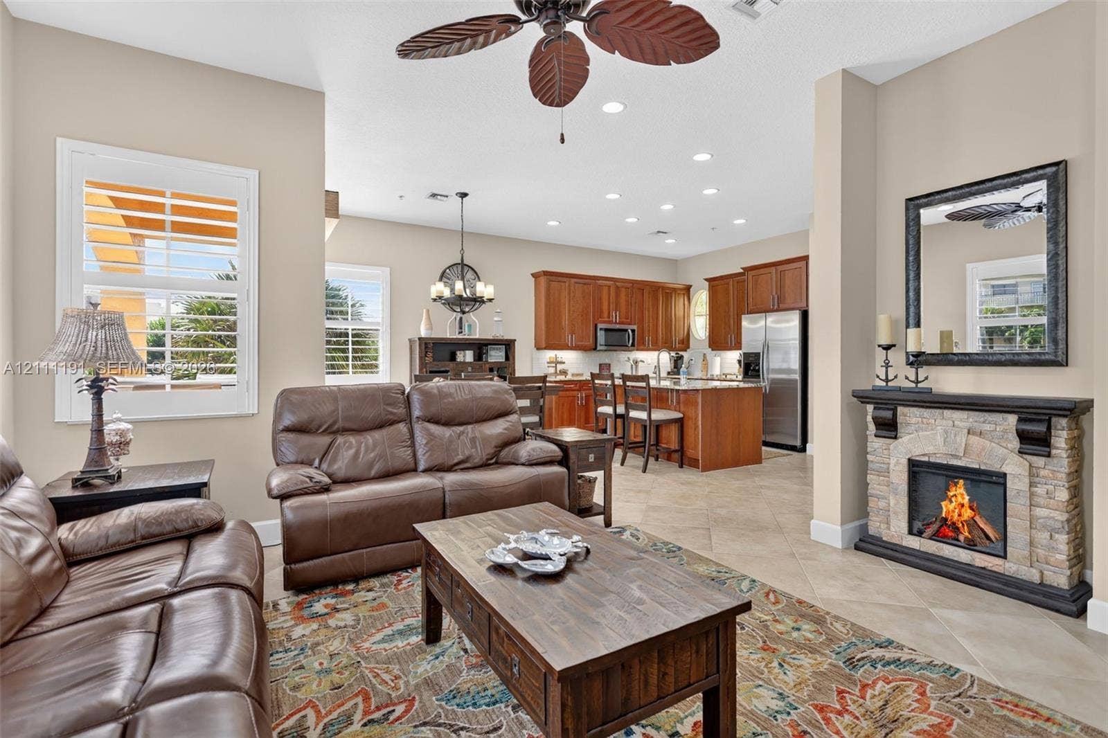 Open-concept living space featuring ceramic tile flooring, stone-veneer fireplace with a hearth mantle, natural wood cabinetry, recessed lighting, and plantation shutters