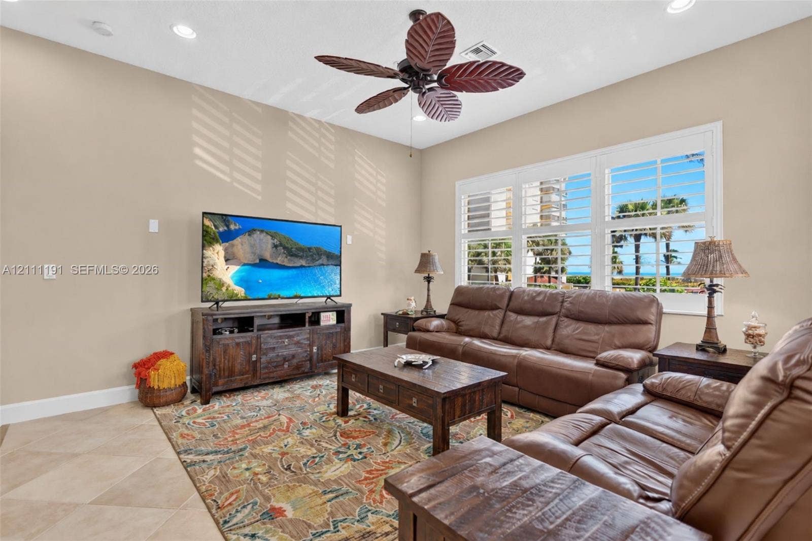 Living area featuring large-format tile flooring, 65" TV, recessed lighting, a decorative ceiling fan with leaf-inspired blades, and expansive windows fitted with plantation shutters