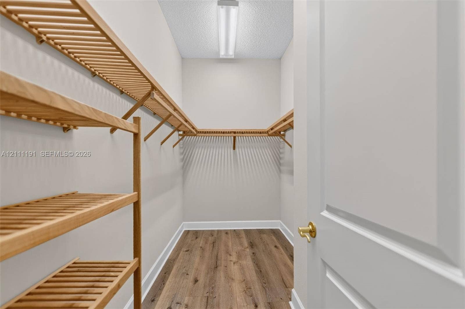 Primary Walk-in Closet. Storage space featuring wood-finish shelving and wood-finish flooring, complemented by a flush-mount overhead light fixture and a panel door with brass-finish hardware
