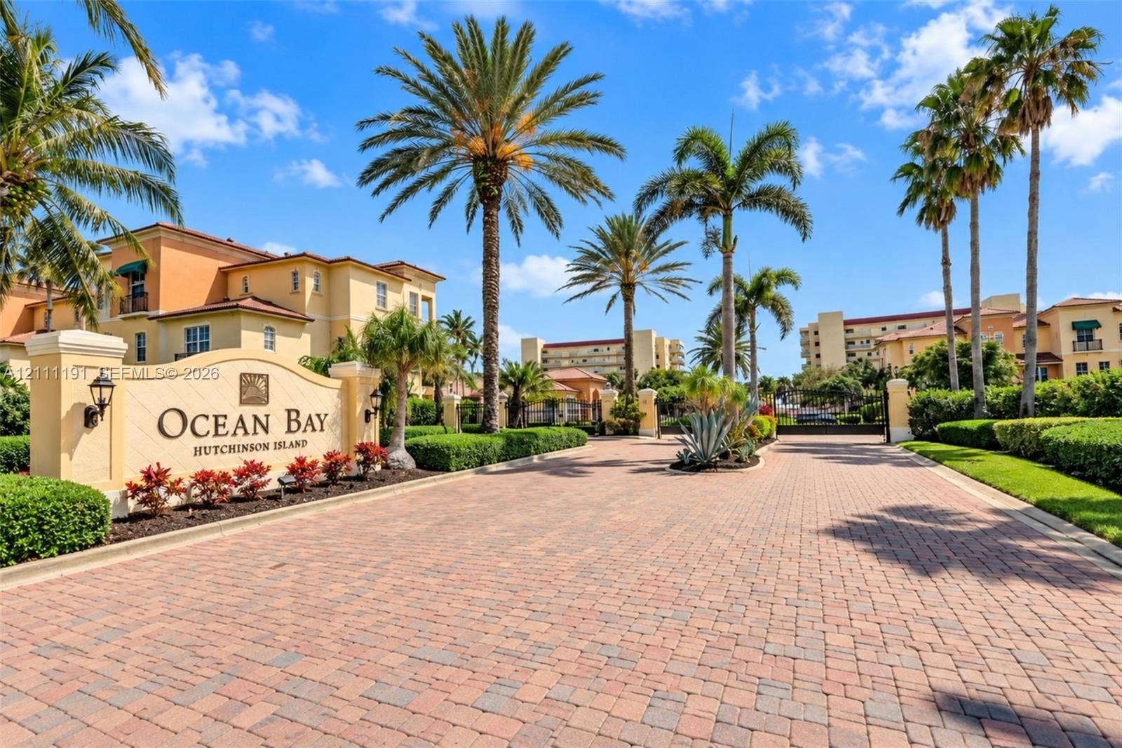 Gated entrance featuring a brick-paver drive, custom stone monument signage, mature palm landscaping, and integrated wall-mounted lighting