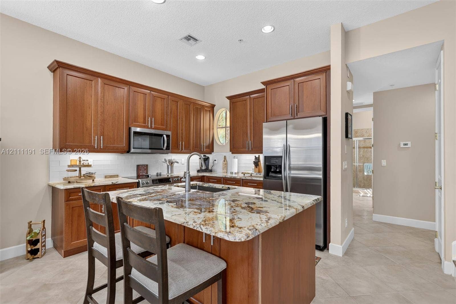 Kitchen featuring natural wood cabinetry, stone-surface countertops, center island with an integrated sink, white tile backsplash, and stainless steel appliances