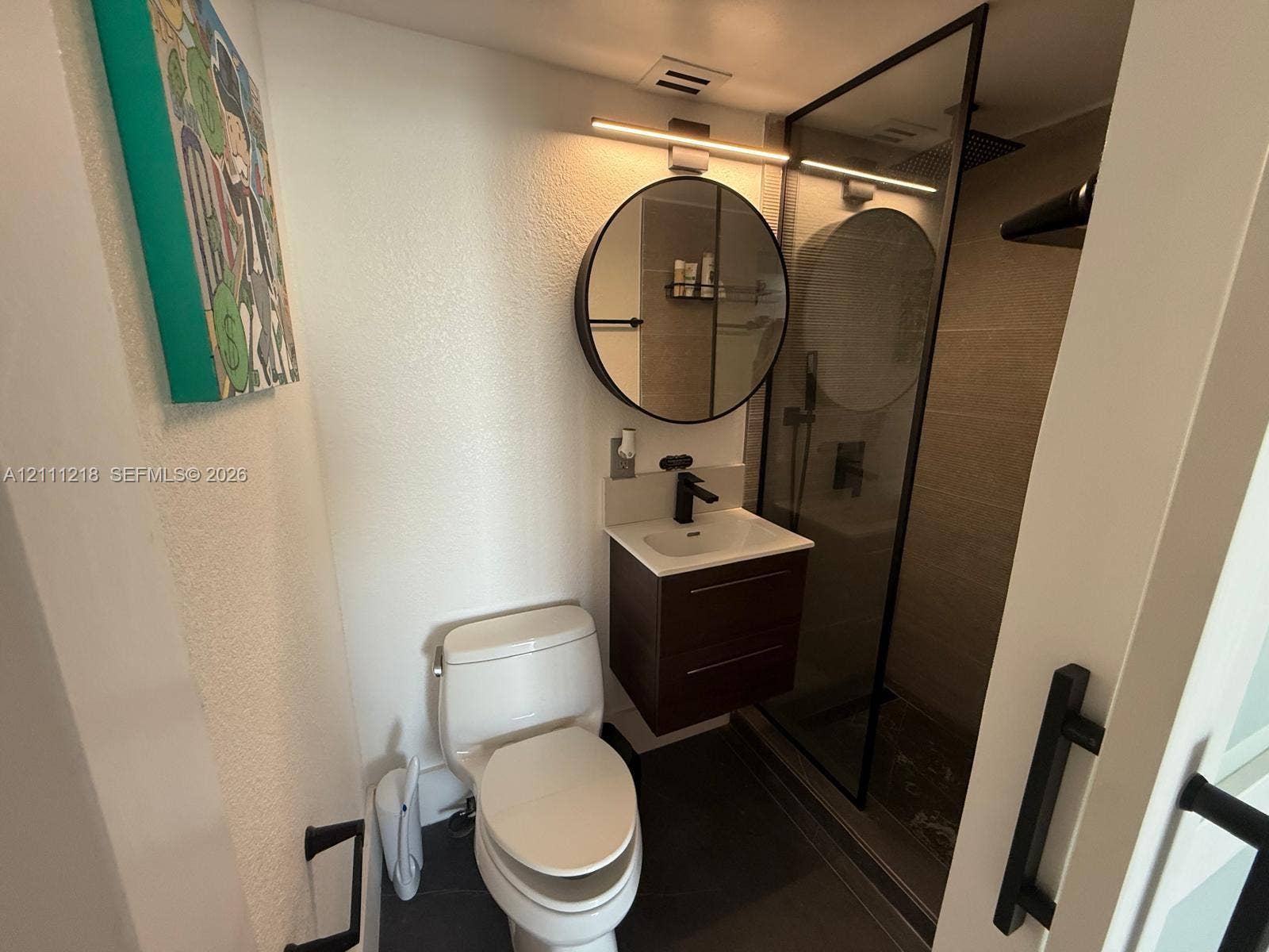 Modern floating vanity with a light-toned countertop and matte black faucet, paired with a circular mirror, linear vanity lighting, and a glass-enclosed shower with tiled surround