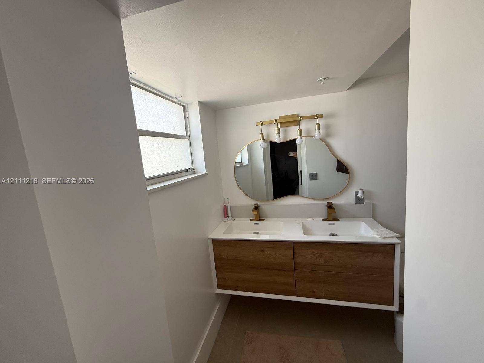 Floating wood-finish vanity with dual integrated sinks, metallic hardware, an organic-shaped wall mirror, and modern multi-bulb lighting