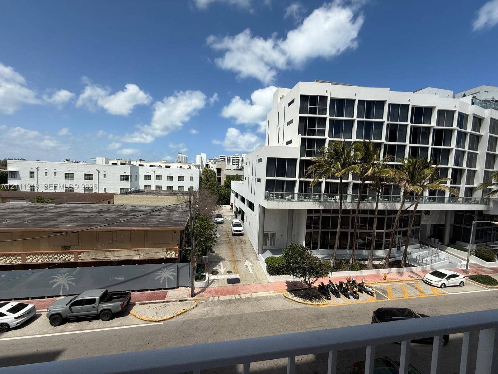 Elevated balcony featuring a white railing, overlooking an urban streetscape with multi-story buildings, palm trees, and clear sky