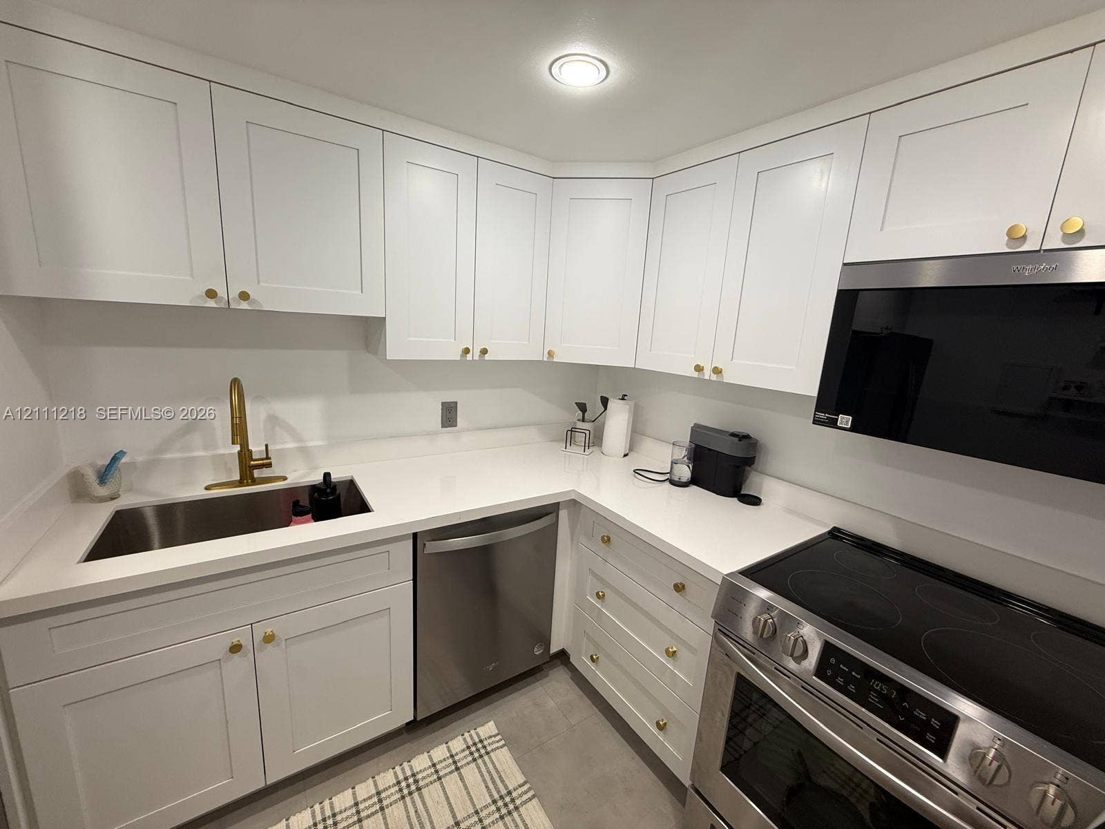 Kitchen featuring white shaker-style cabinetry, solid-surface countertops, stainless steel appliances, and brass-tone hardware