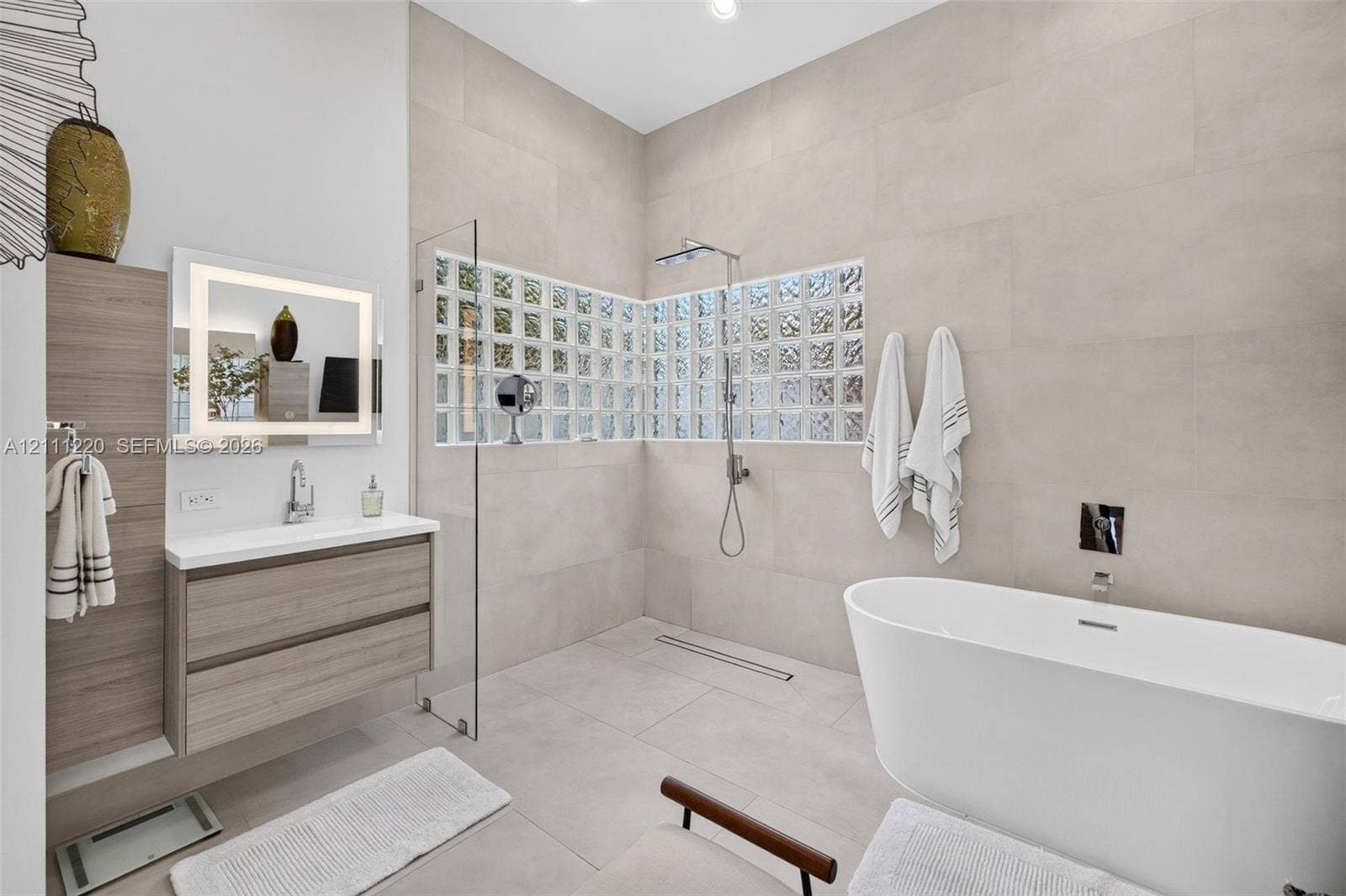 134 Dockside Terrace, Weston, FL 33327 Bathroom featuring a freestanding soaking tub, walk-in shower with glass partition, neutral large-format wall tile, wood-finish wall-mounted vanity, and glass block accent windows