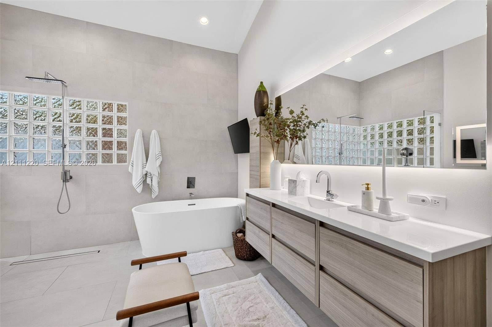 134 Dockside Terrace, Weston, FL 33327 Bathroom featuring a floating wood-finish vanity with a countertop basin, a standalone soaking tub, full-wall tiling, a glass-block privacy window, and an expansive backlit mirror
