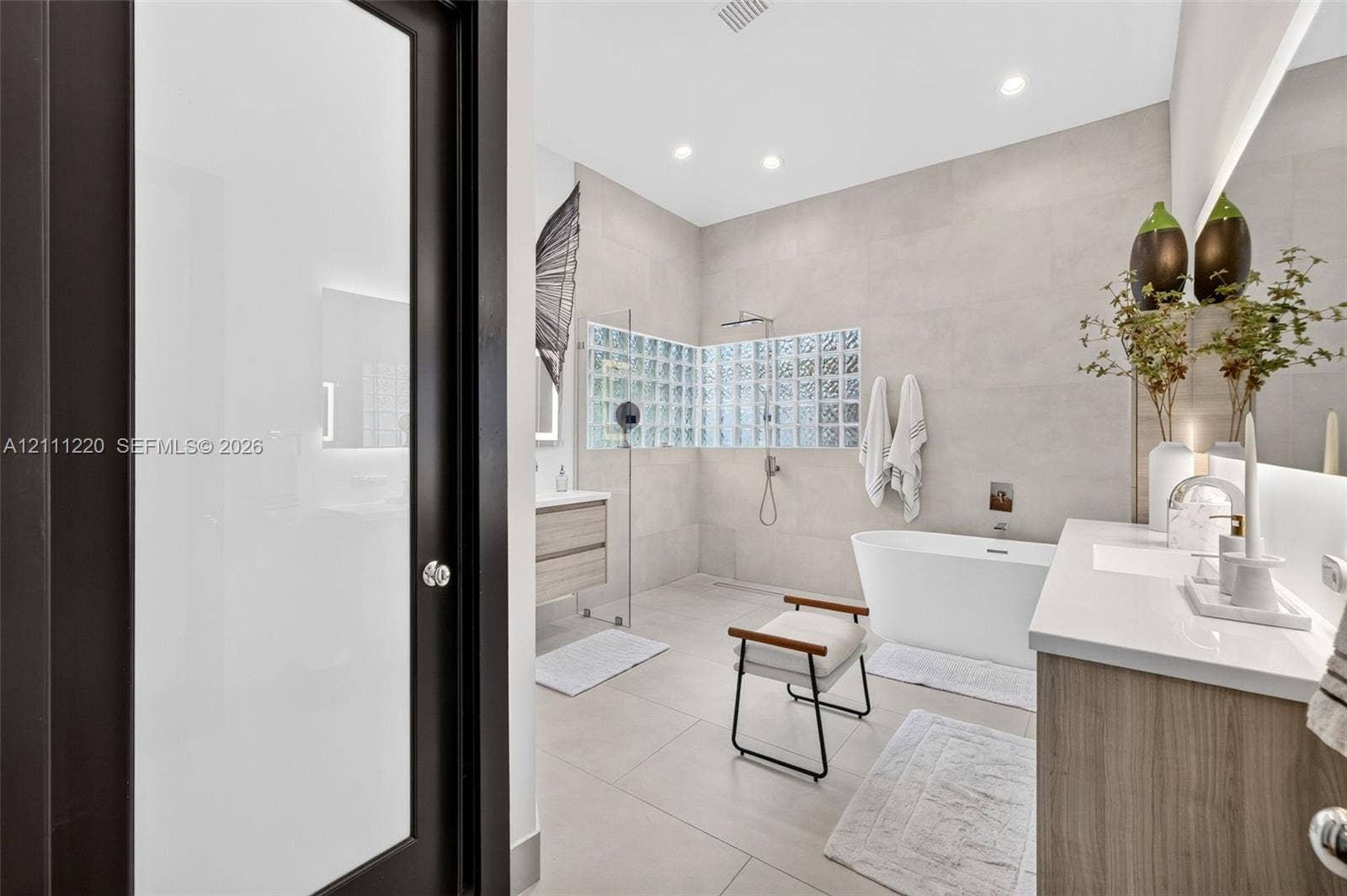 134 Dockside Terrace, Weston, FL 33327 Bathroom featuring large-format tile flooring and walls, a freestanding soaking tub, a wet-room style shower enclosure with glass block wall detailing, and a wood-finish vanity with an integrated countertop