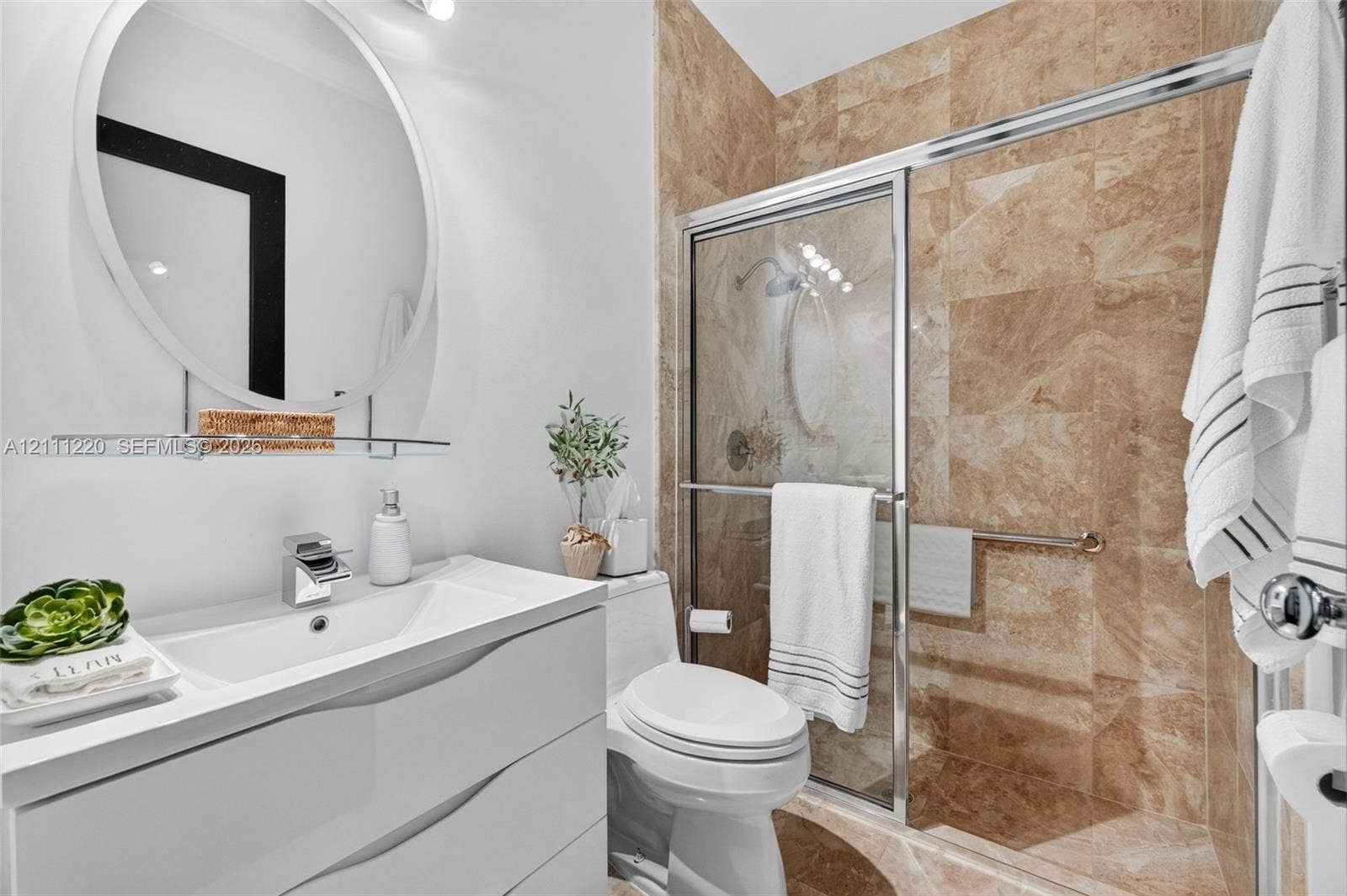 134 Dockside Terrace, Weston, FL 33327 Contemporary white vanity with integrated basin, polished chrome faucet, circular wall mirror, and walk-in shower featuring floor-to-ceiling stone-look tiling and sliding glass enclosure